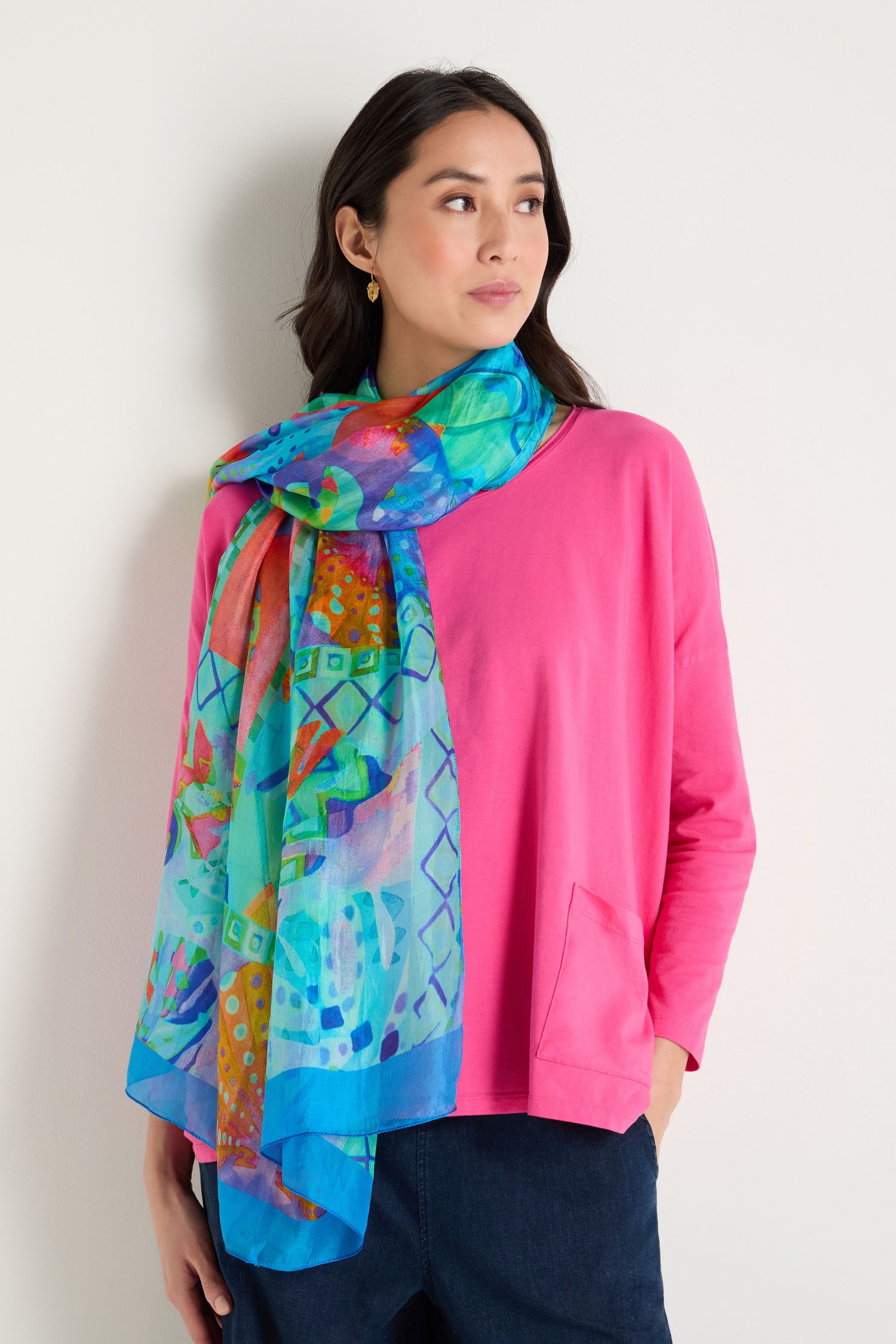 A woman in a bright pink long-sleeve top and dark pants stands against a white wall, accessorized with the Ocean Garden Silk Scarf draped around her neck, featuring a vibrant coastal print and luxurious sheen.