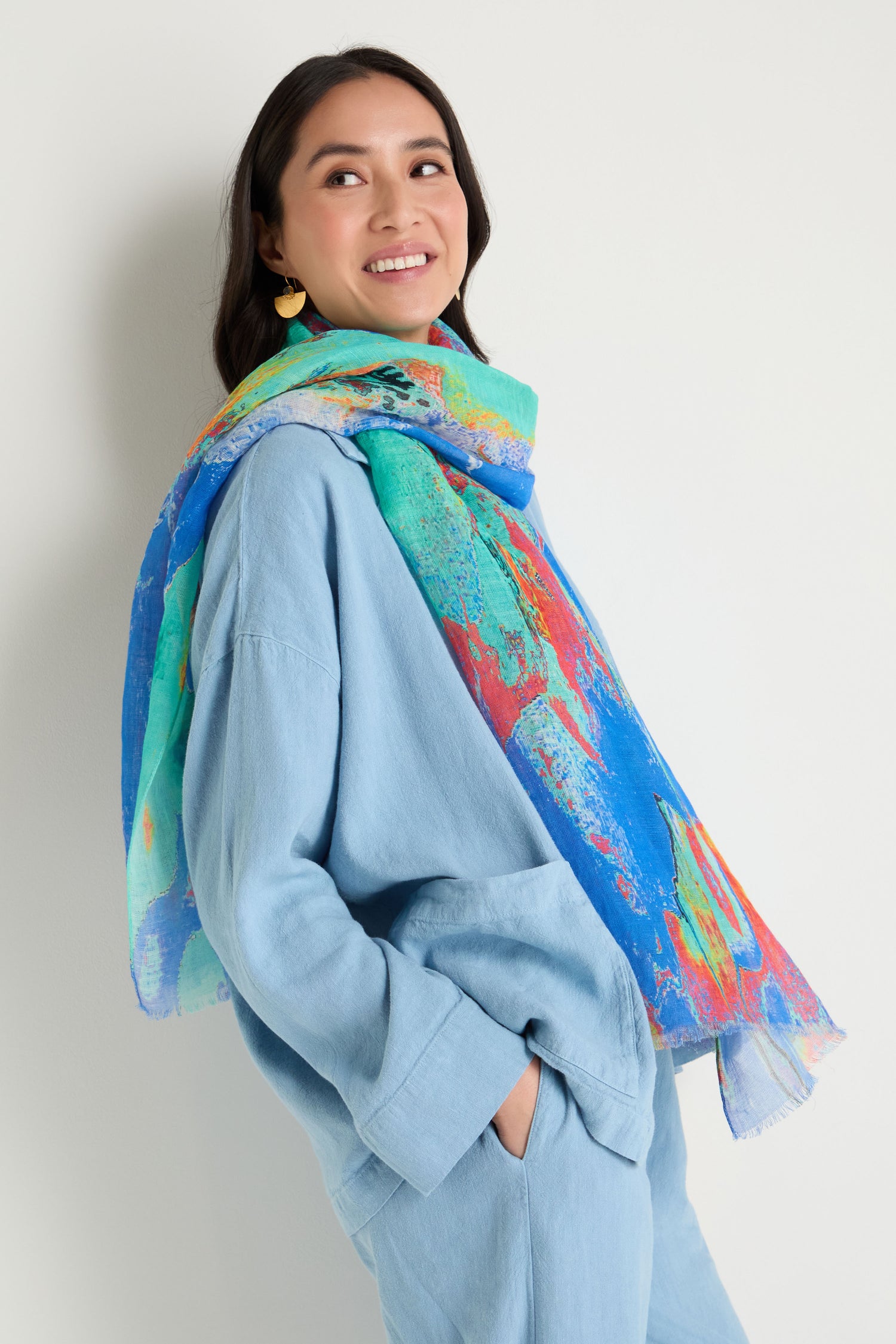 A woman smiles and looks to the side, standing with hands in pockets against a plain background, wearing a light blue outfit paired with the Terra Mare Cotton Linen Scarf.