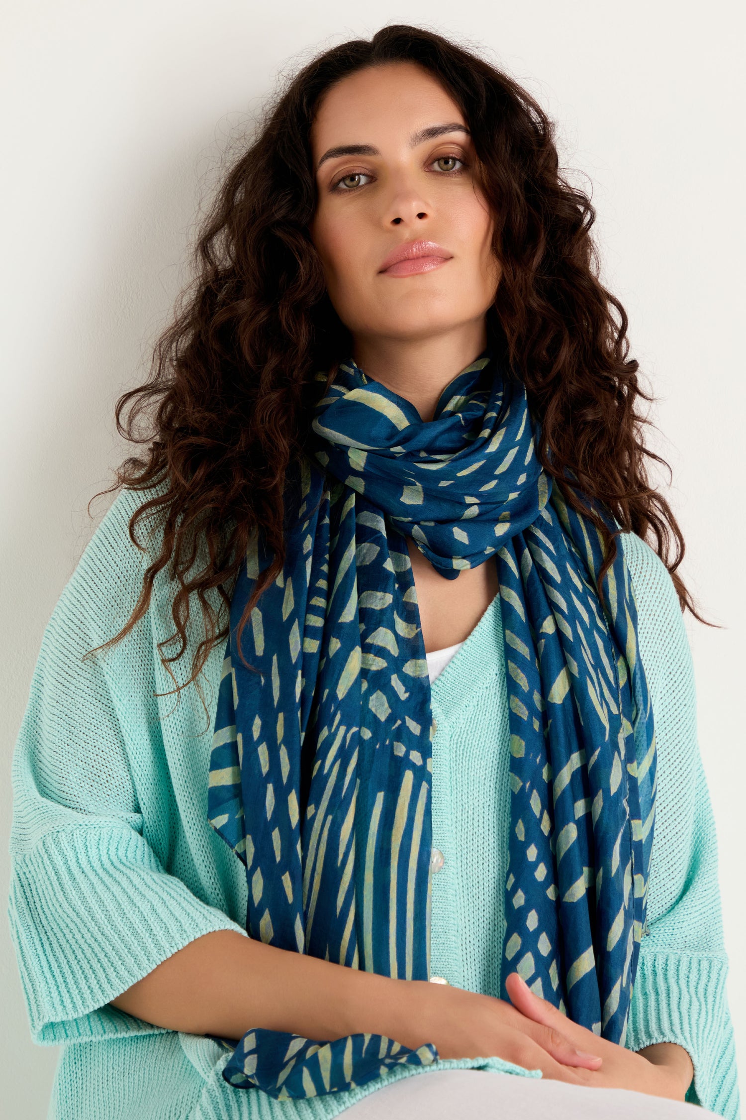 A woman with long curly hair wears a light blue top and the Painted Waves Silk Scarf with an abstract print, sitting against a plain white background.