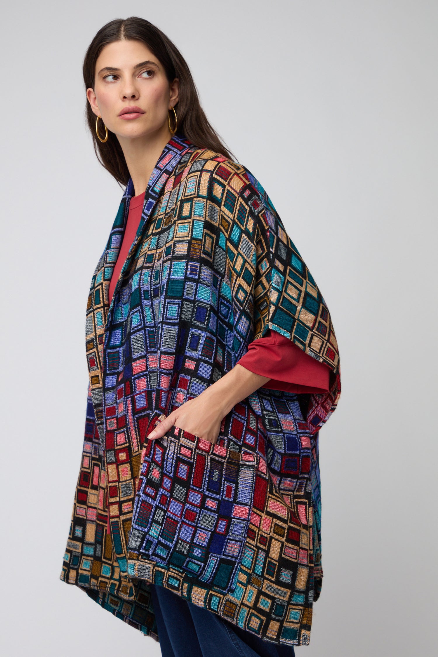 A woman wears the Tesserae Jacket, a contemporary outerwear piece with a multicolored geometric pattern, layered over a red shirt. She stands against a plain light gray background.