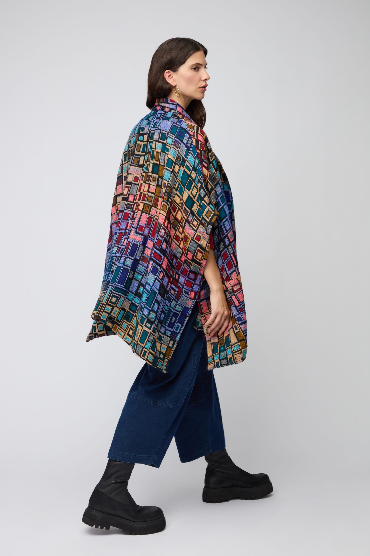 A woman stands in profile against a plain background wearing the vibrant Tesserae Jacket, featuring bold geometric patterns, styled with wide-leg blue pants and black chunky boots.