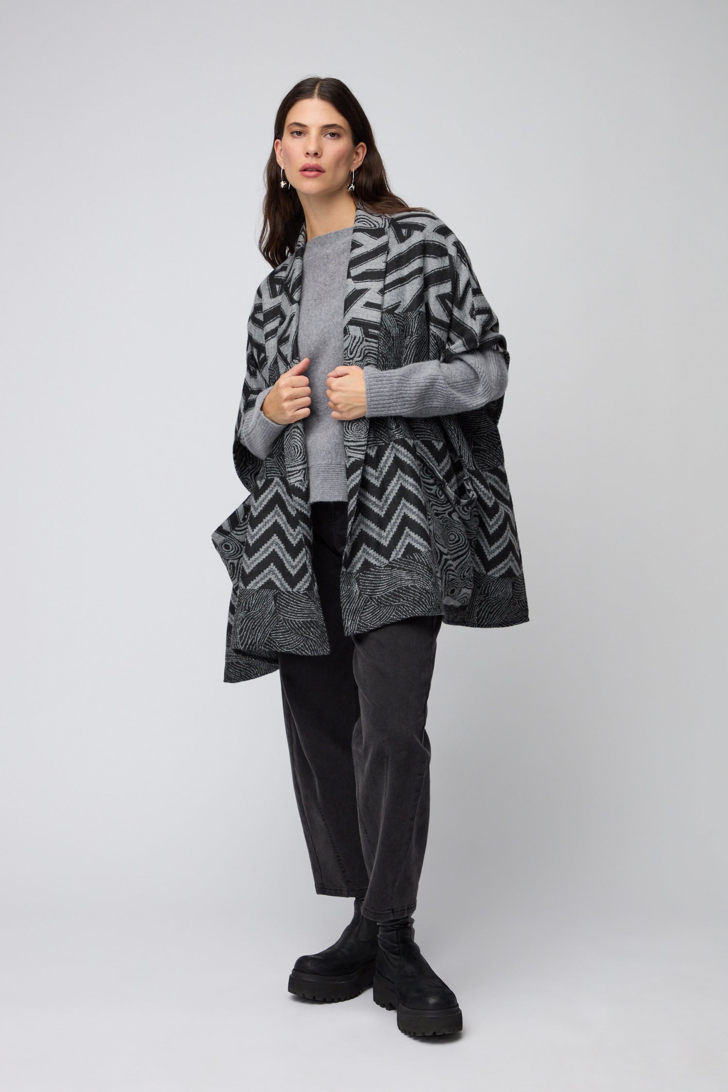 A woman stands wearing the Zig Zag Jacket in black and grey, styled with a grey sweater, black pants, and black boots against a plain light background.