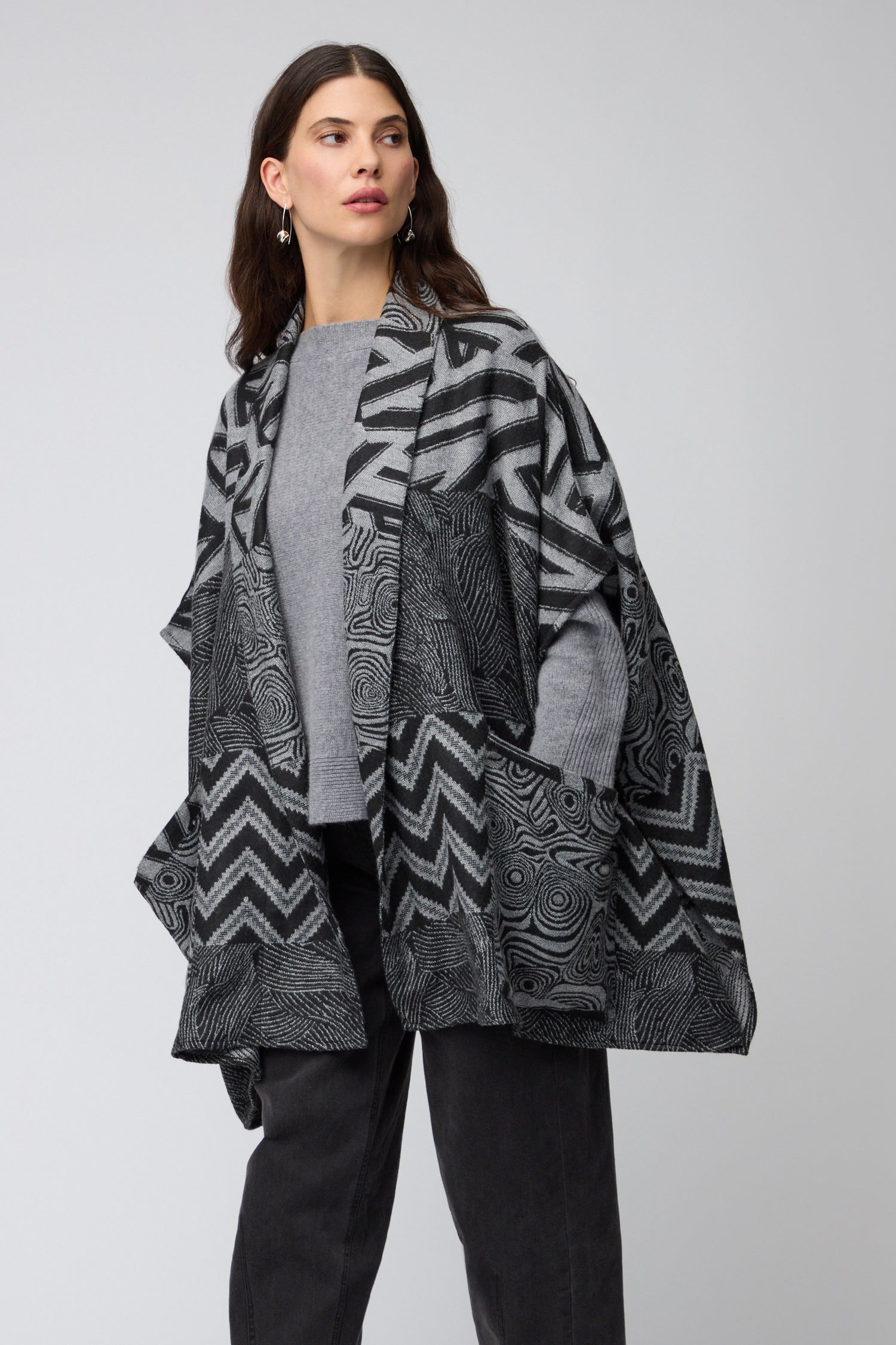 A woman models the Zig Zag Jacket, a versatile black and grey patterned layer, over a grey sweater and black pants, standing against a plain light background.