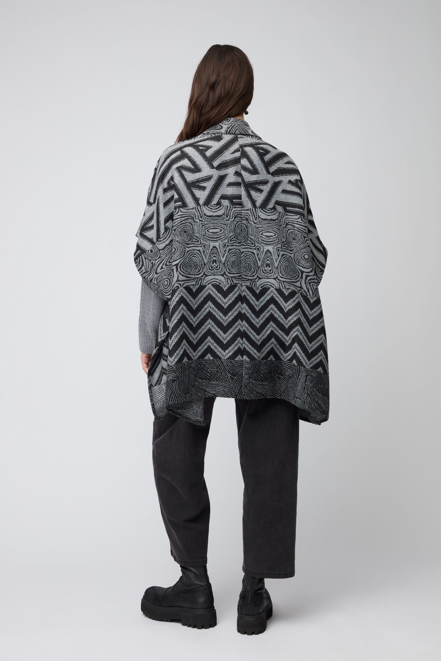 A person faces away, wearing the Zig Zag Jacket—a black and grey patterned layering piece—paired with dark pants and black boots against a plain light background.