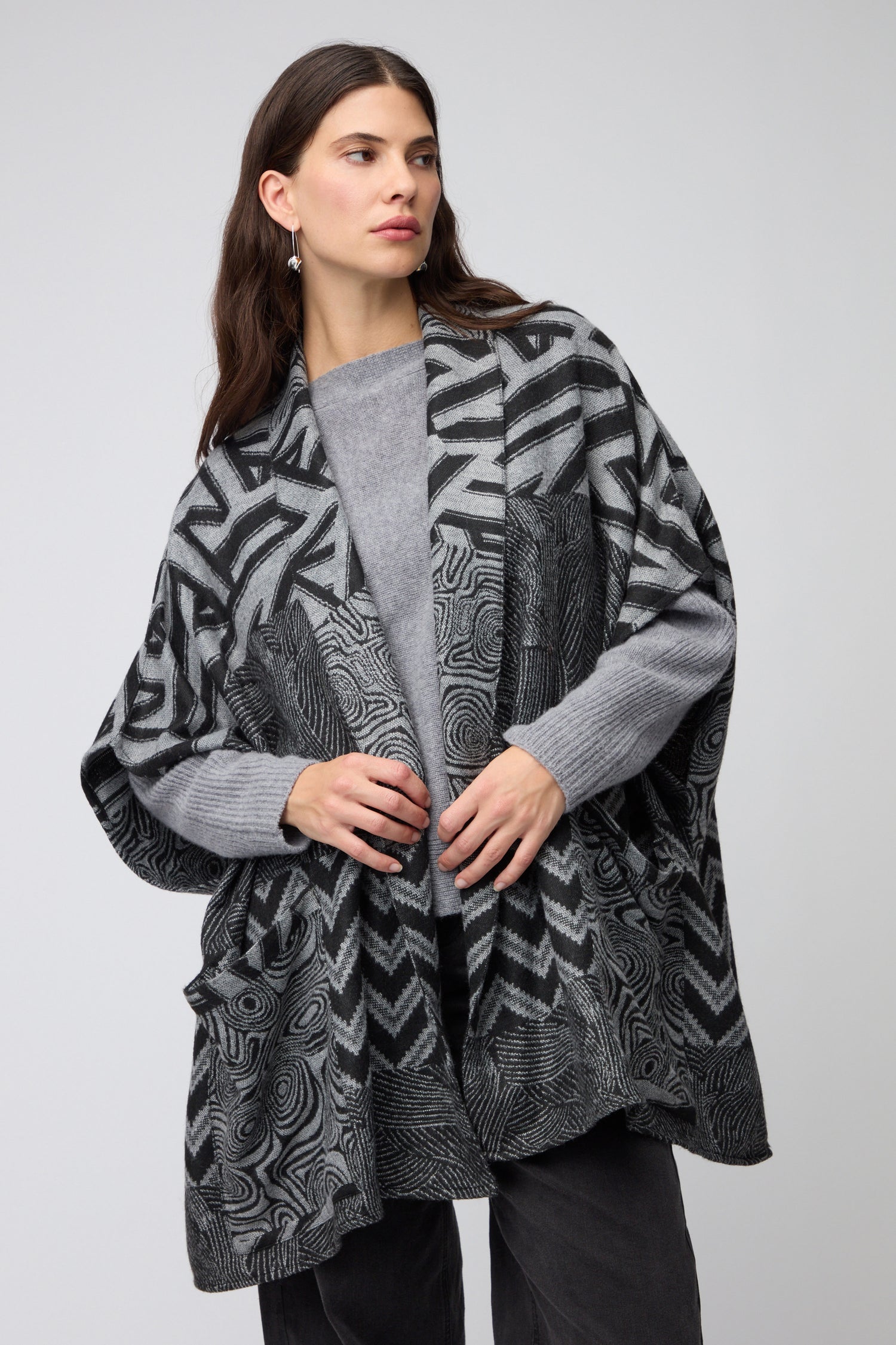 A woman wears the Zig Zag Jacket—a patterned, loose-fitting black and grey layer—over a grey sweater and dark pants, standing against a plain light background.