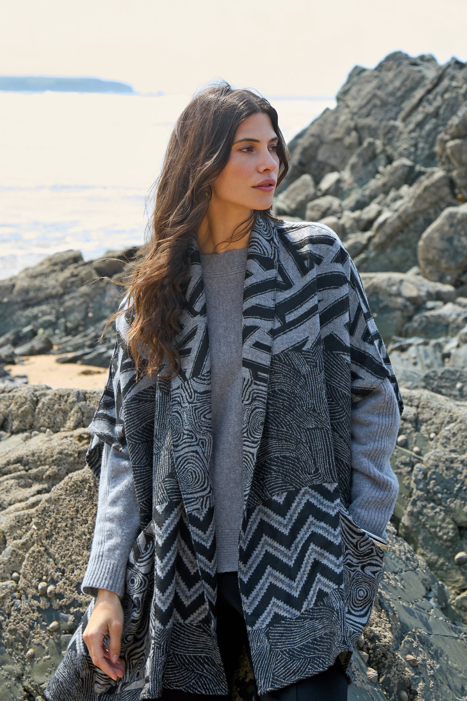 A woman stands on rocky terrain by the sea, wearing a geometric-patterned Zig Zag Jacket over a grey sweater.