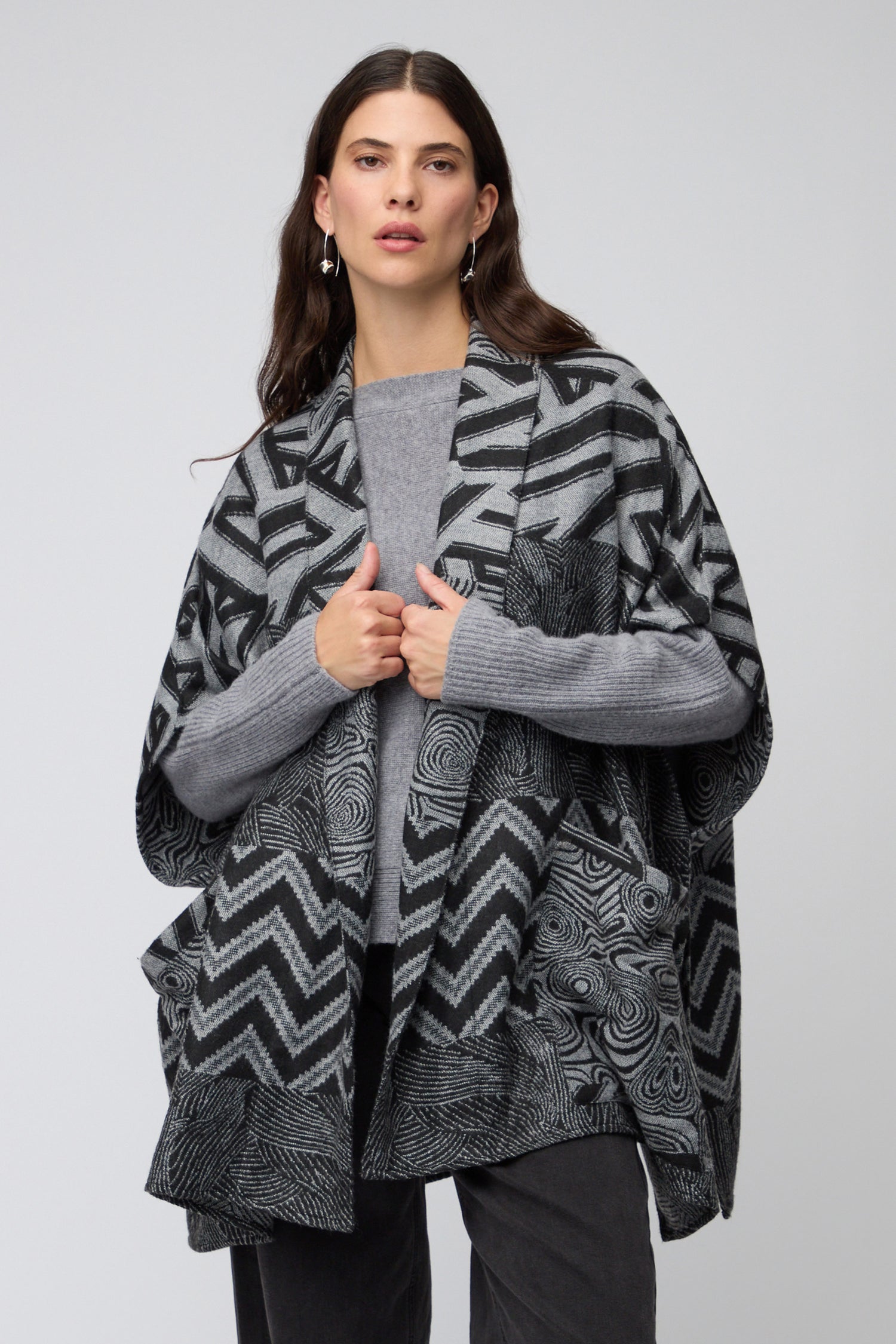 Model wearing the Zig Zag Jacket, a grey patterned shawl-style layer, over a grey sweater and dark pants, posed against a plain studio background.
