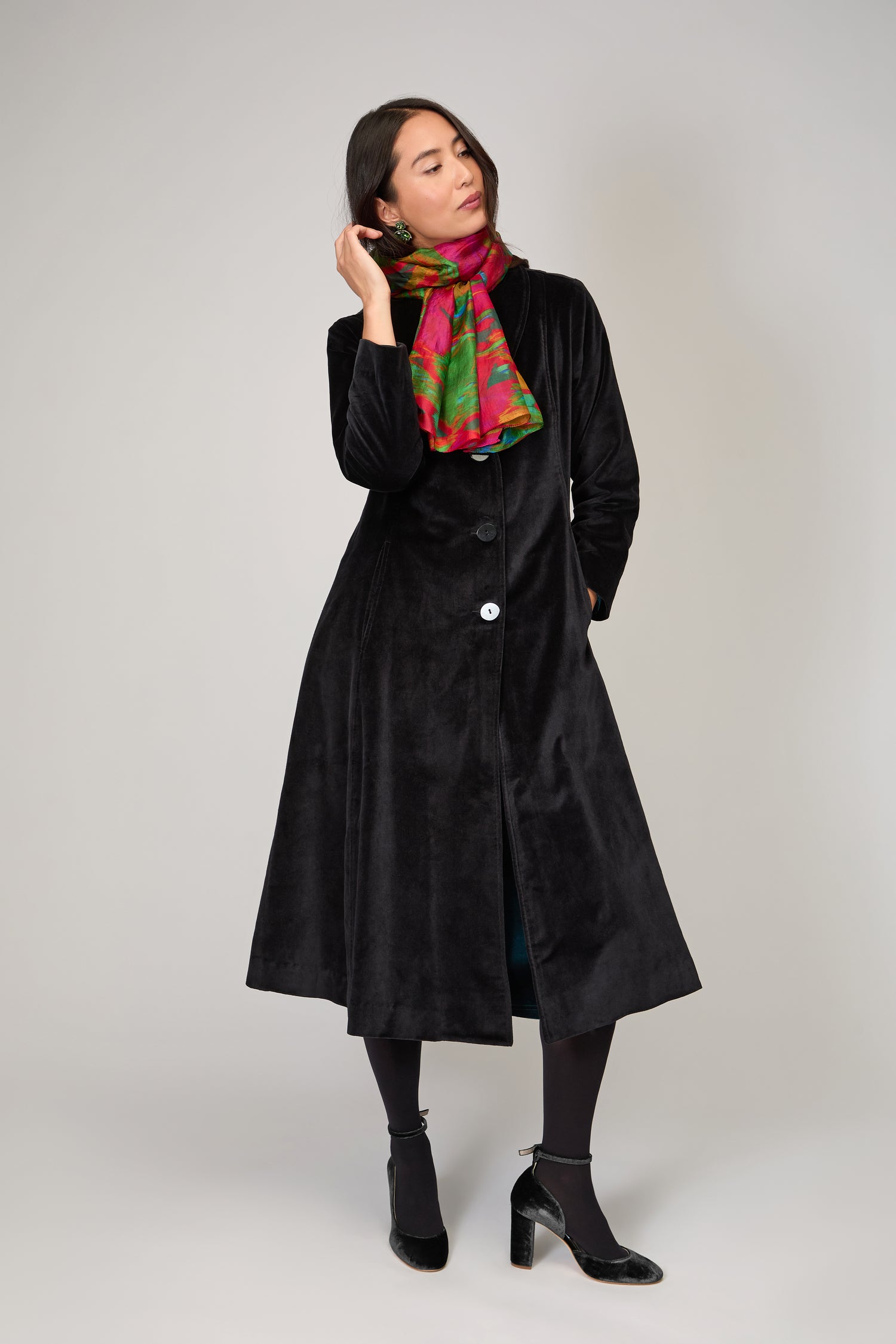 A woman stands against a plain light background, styled in a long black velvet coat, black tights, black heels, and accessorized with the Blurred Botanicals Scarf.