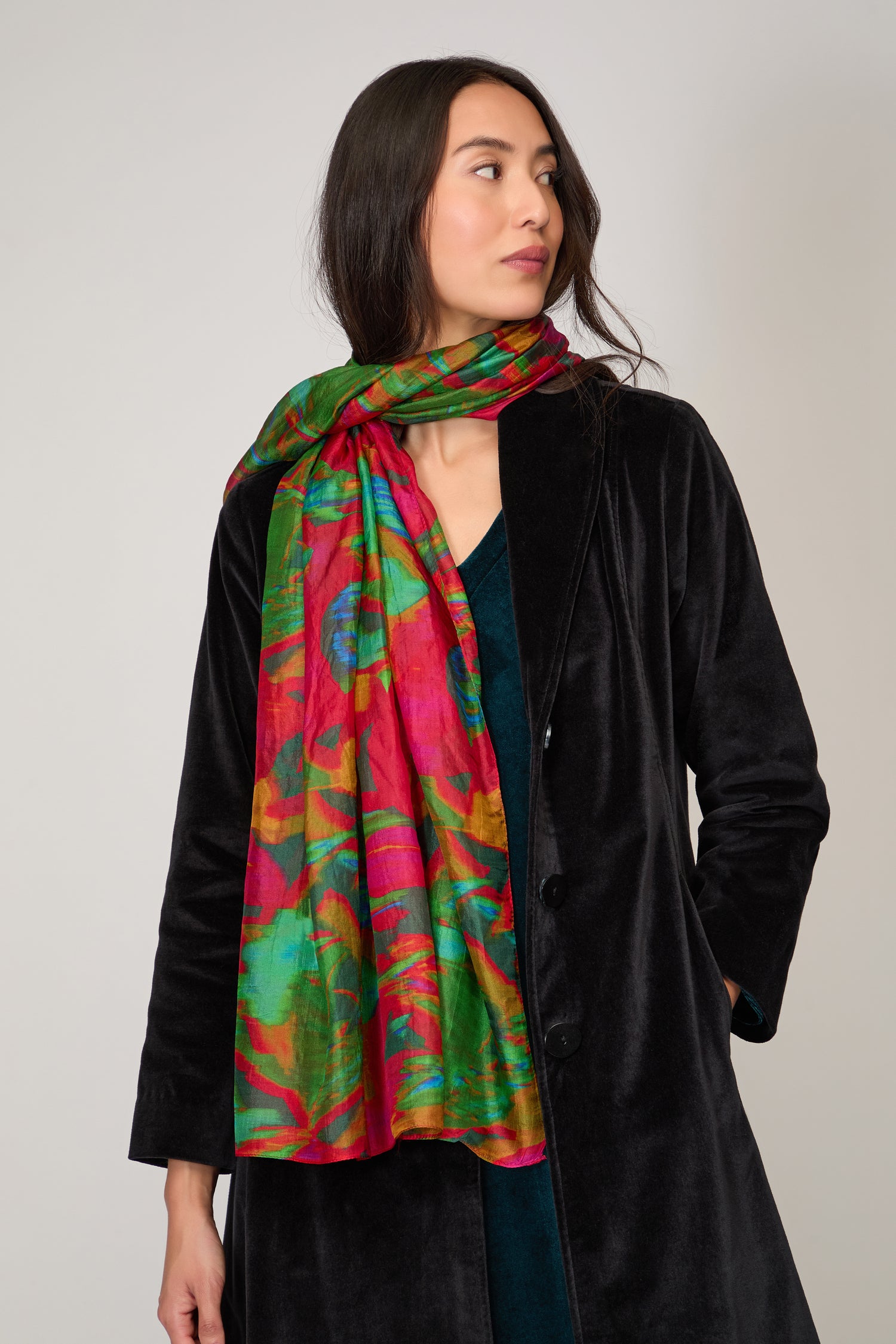 A person in a black coat styles their look with the Blurred Botanicals Scarf, standing against a plain light background and gazing to the side.
