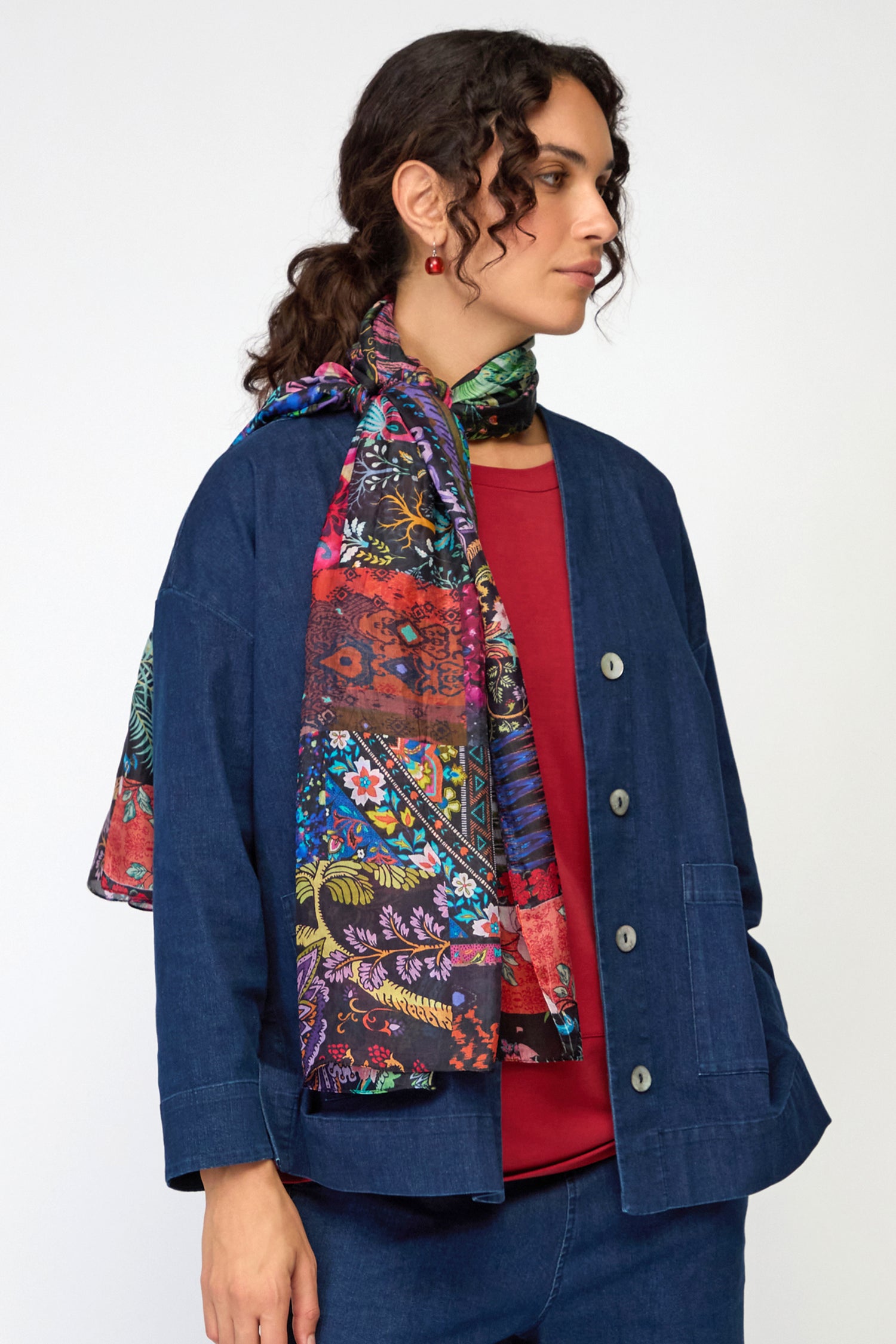 A woman wearing a blue denim jacket and pants, a red top, and the Sahara Winter Patchwork Scarf stands against a plain light background.