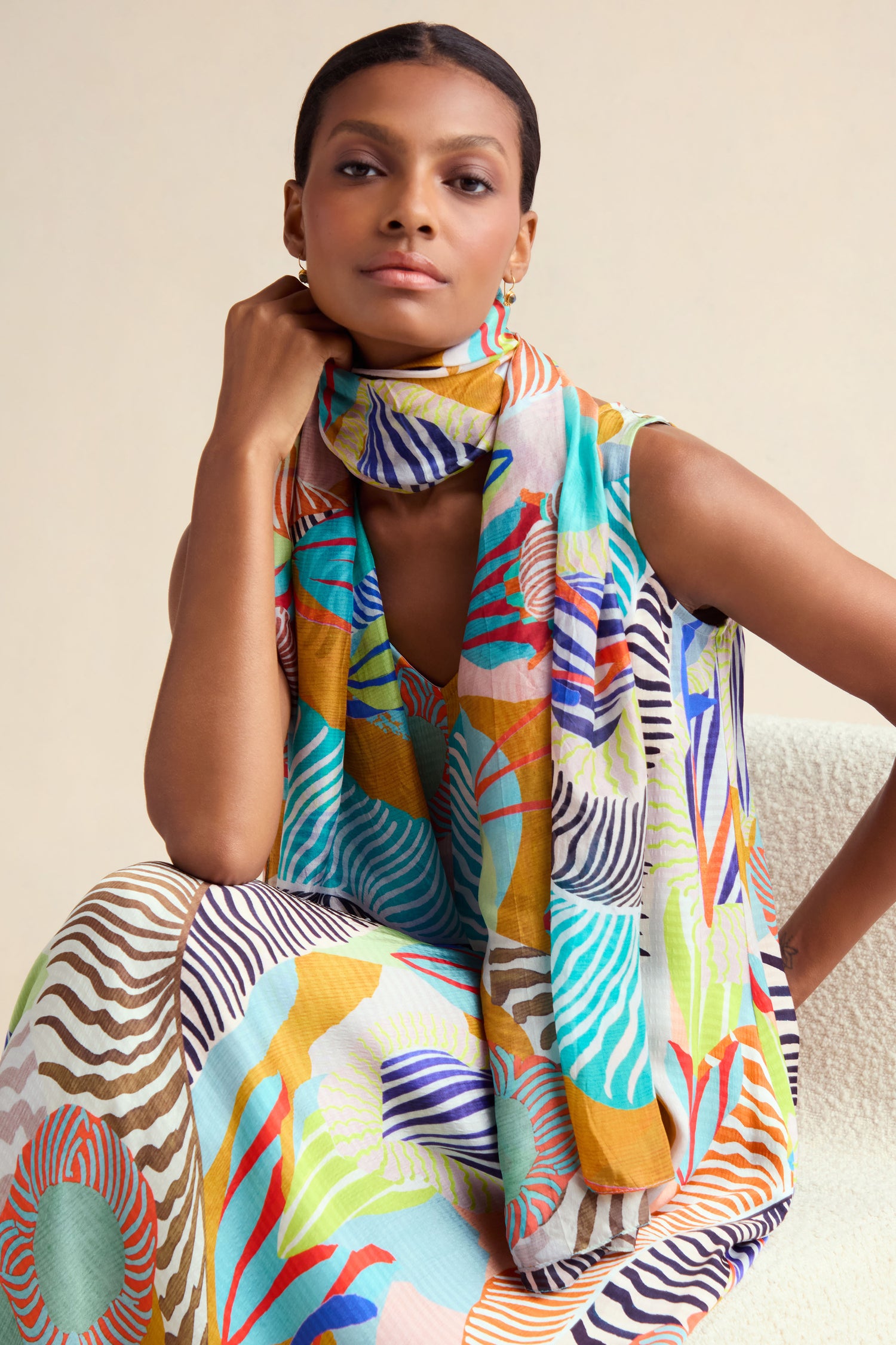 A person wearing a sleeveless dress and the Tropical Orchid Fantasy Silk Scarf, featuring a colorful abstract pattern, sits on a chair with one hand resting on their chin.