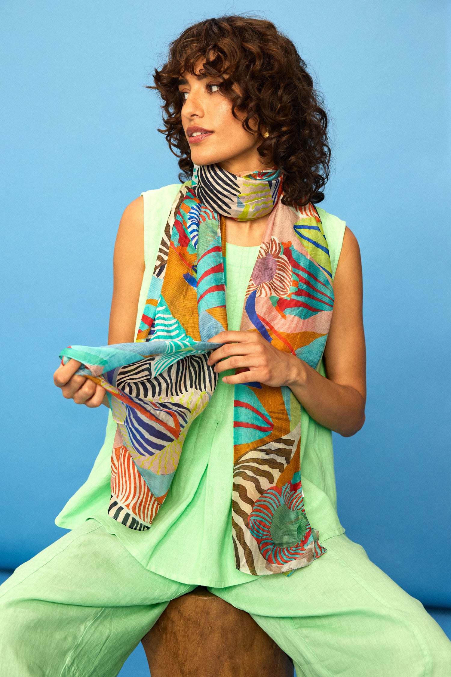 A woman with curly hair wears a light green outfit and the Tropical Orchid Fantasy Silk Scarf while seated on a wooden stool against a blue backdrop.