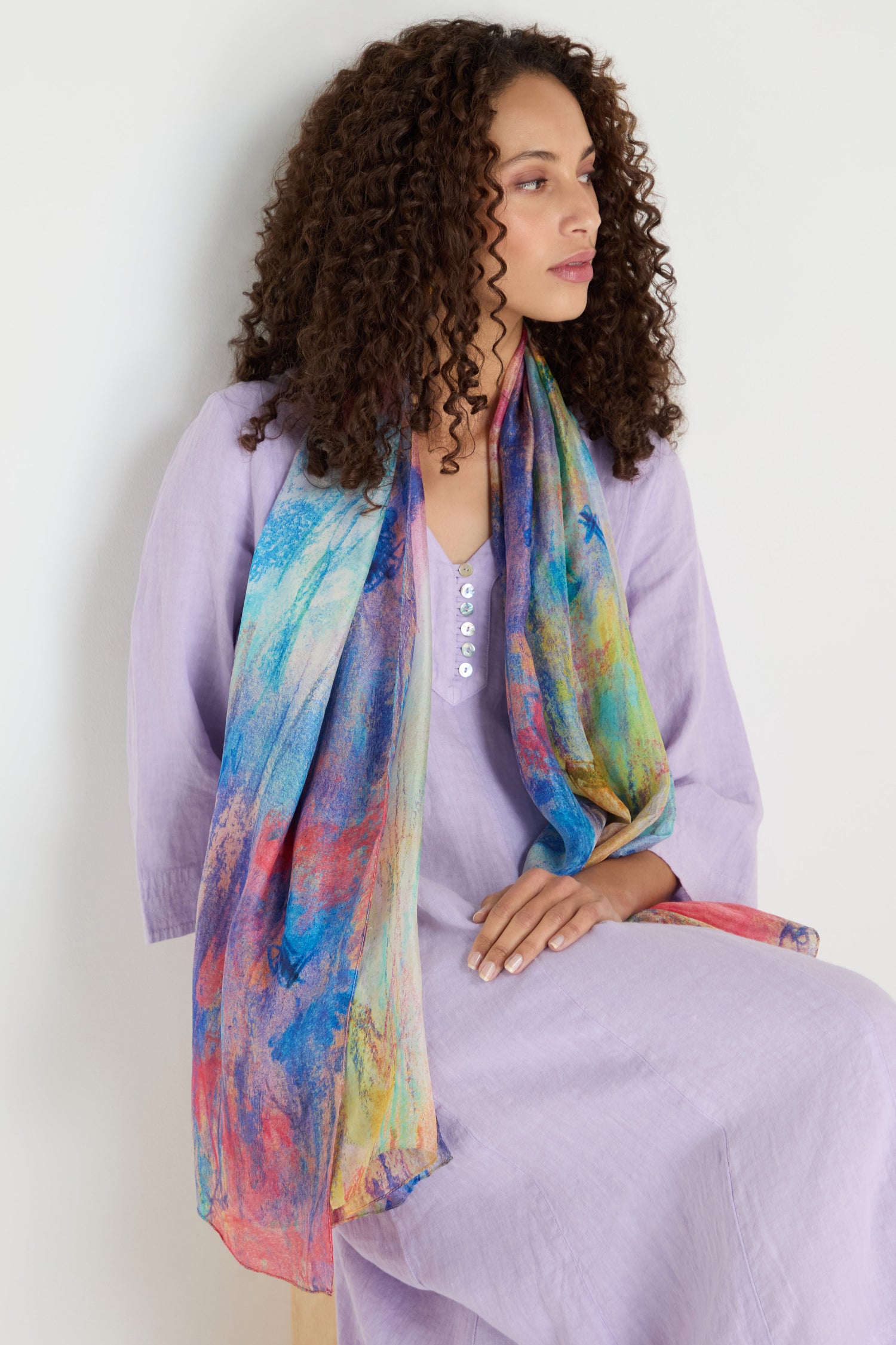 A woman with curly hair, in a light purple dress and the Soft Meadow Dreams Silk Scarf, sits on a stool against a plain white background.