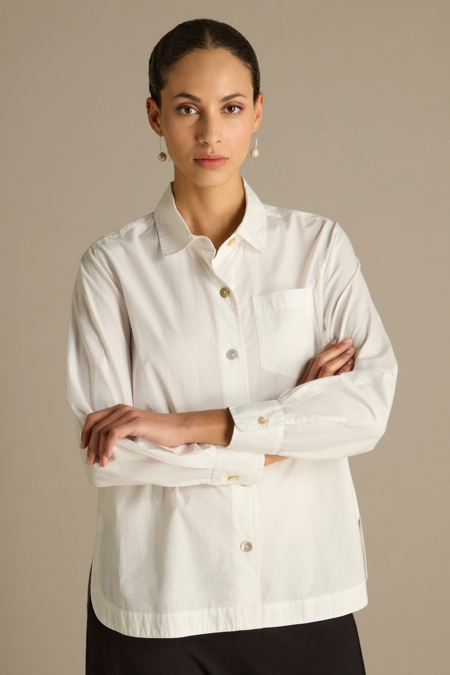 A person with dark hair stands confidently, arms crossed, wearing the Curved Hem Shirt—a white button-up crafted from premium cotton.