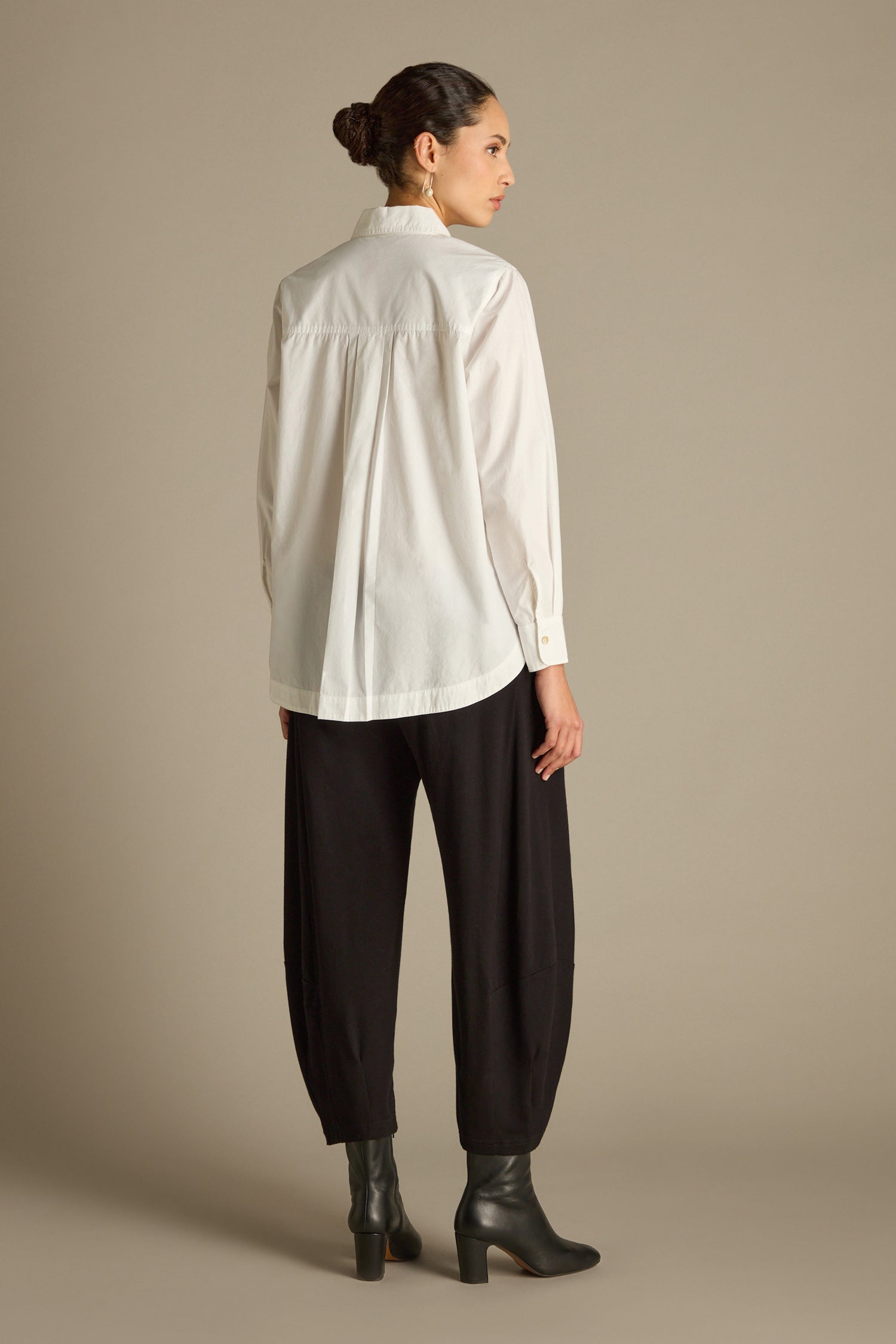 A person with tied-back hair wearing a white curved hem shirt made of Indian cotton, paired with black pants and heeled boots, stands facing away in a neutral pose against a plain background.