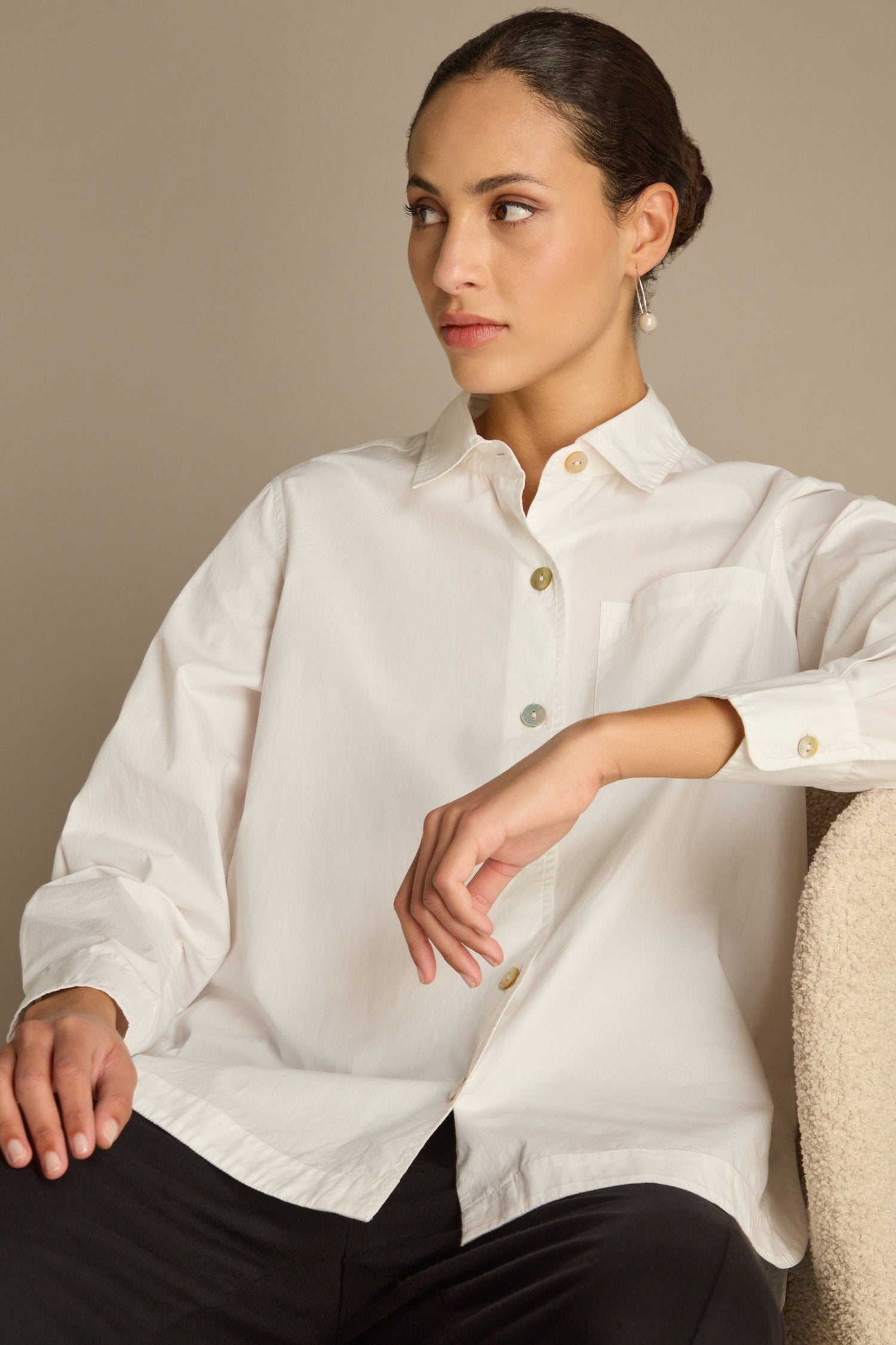 Individual clad in a Curved Hem Shirt made of premium cotton, sitting on a chair against a beige backdrop, glancing to the side, teamed with black pants.