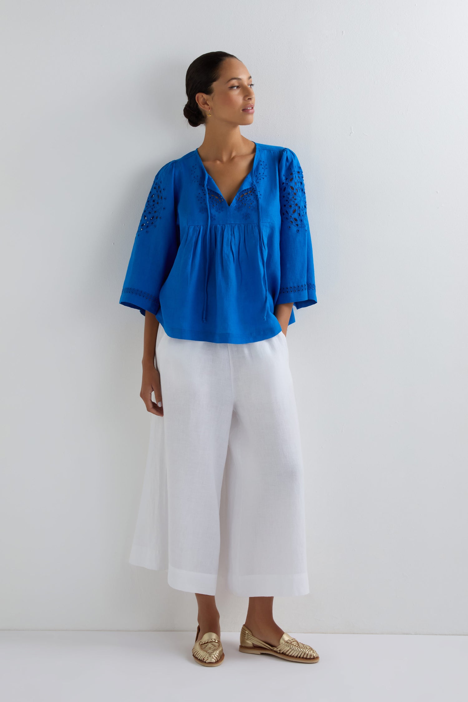 A woman stands against a white wall wearing the Embroidered Schiffli Linen Top with 3/4 sleeves, paired with white wide-leg pants and gold loafers. The breathable linen keeps her comfortable, while delicate embroidery adds sophistication.