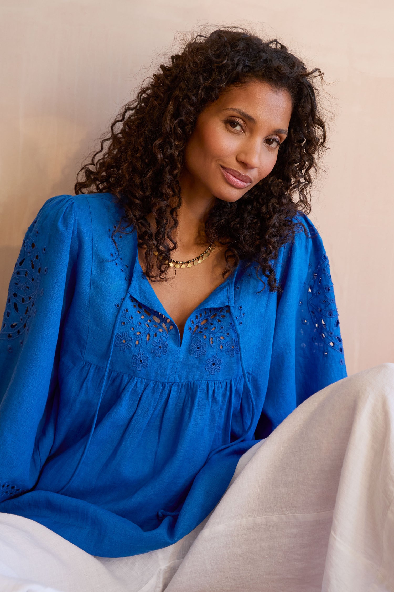 Woman with curly hair wearing a bright blue Embroidered Schiffli Linen Top and white pants, sitting against a neutral background.