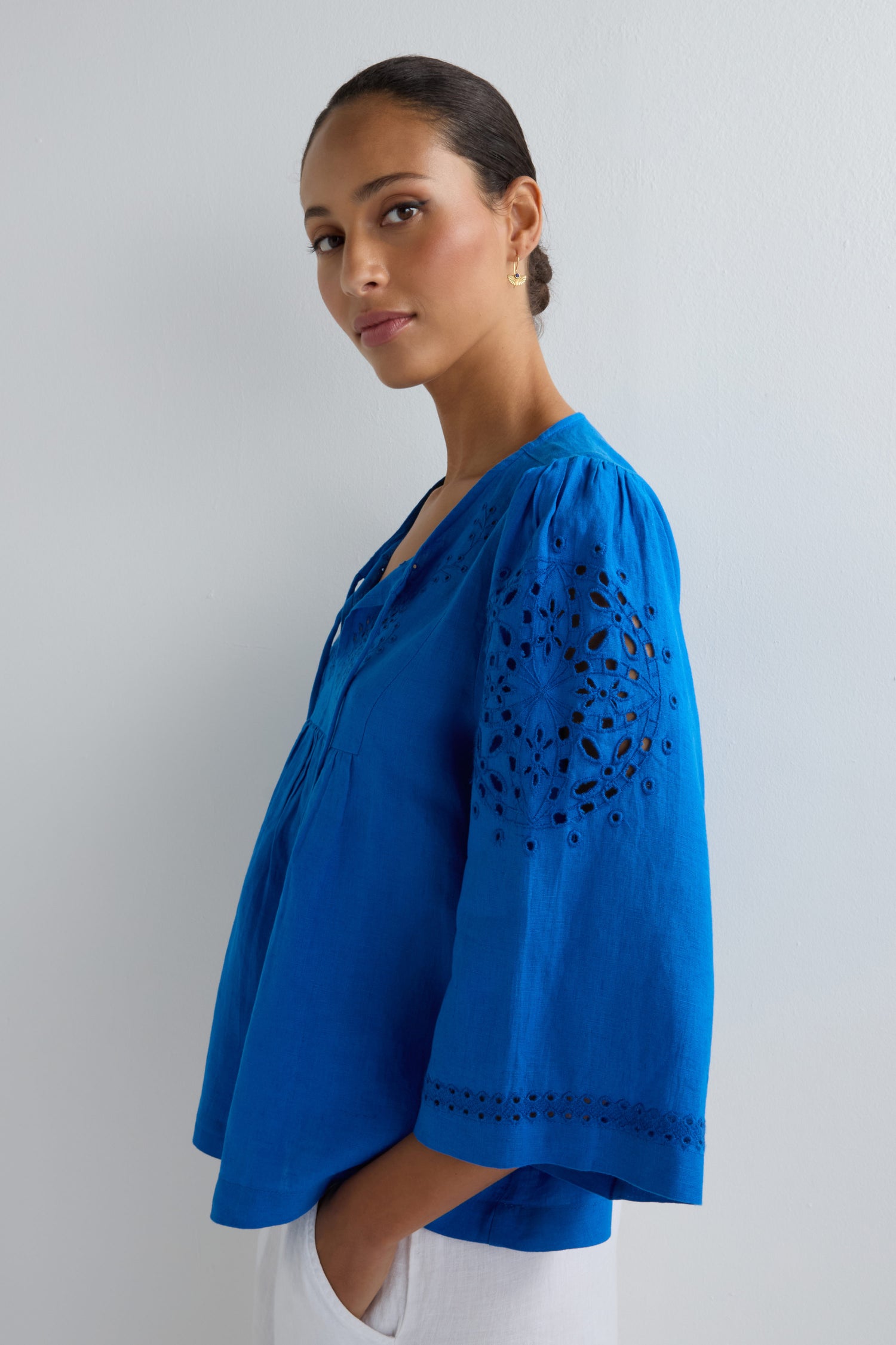 A woman stands sideways in a bright blue Embroidered Schiffli Linen Top with eyelet details and 3/4 sleeves, paired with white pants, set against a plain light background.