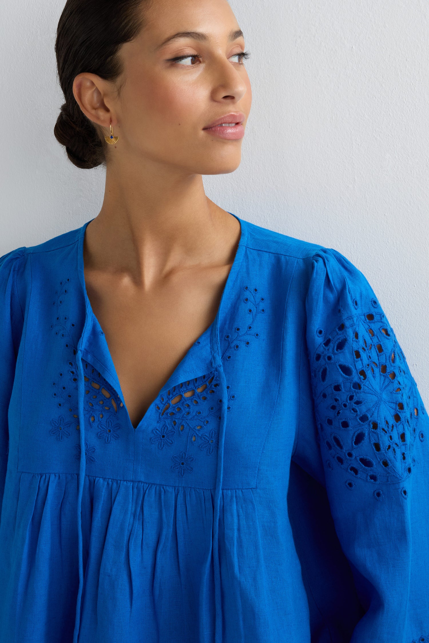 A woman with dark hair in a low bun wears the Embroidered Schiffli Linen Top, featuring 3/4 sleeves and cut-out floral details, while looking to the right against a plain white background.