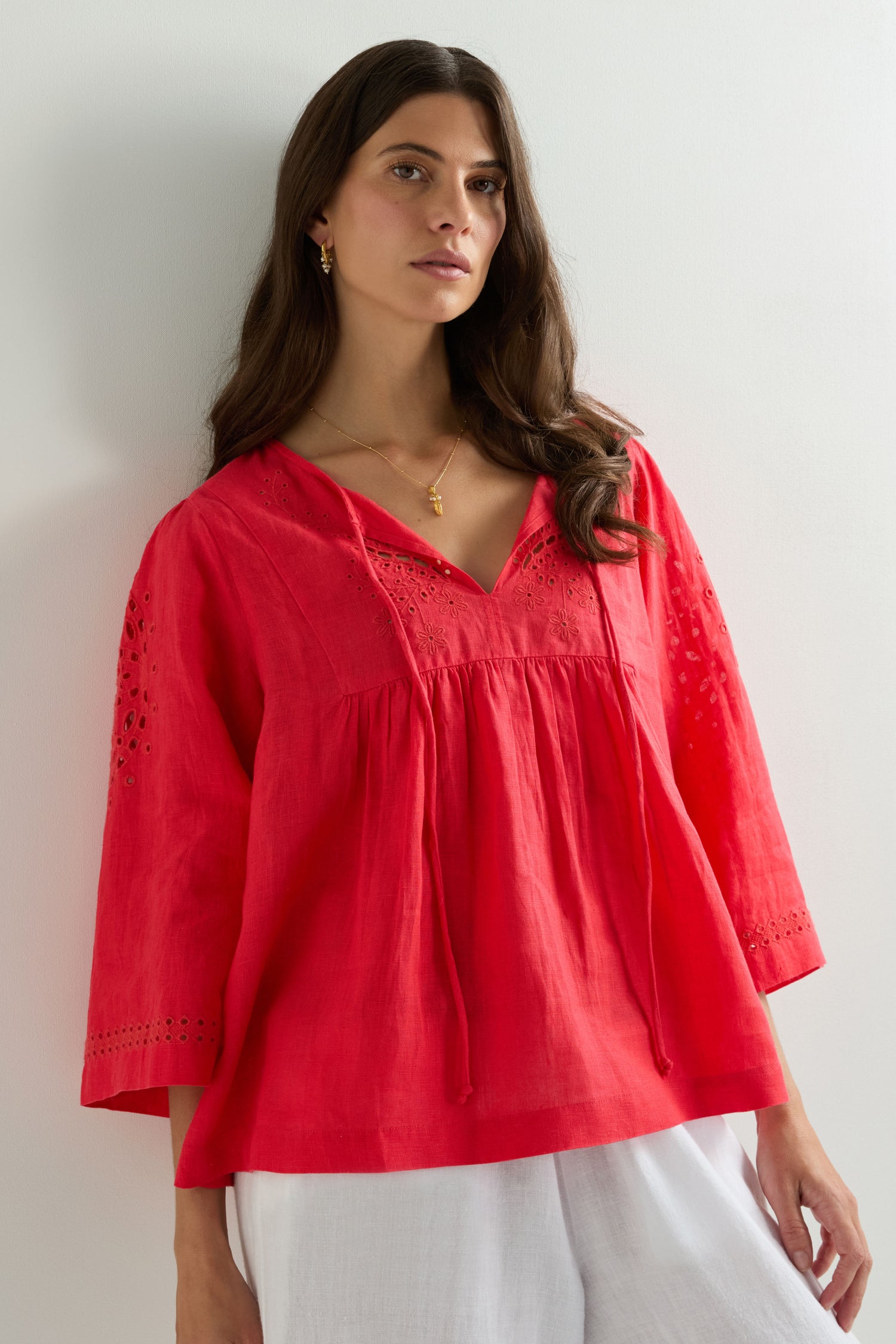 A woman in a loose red Embroidered Schiffli Linen Top with eyelet sleeve details and white pants stands against a plain white background.