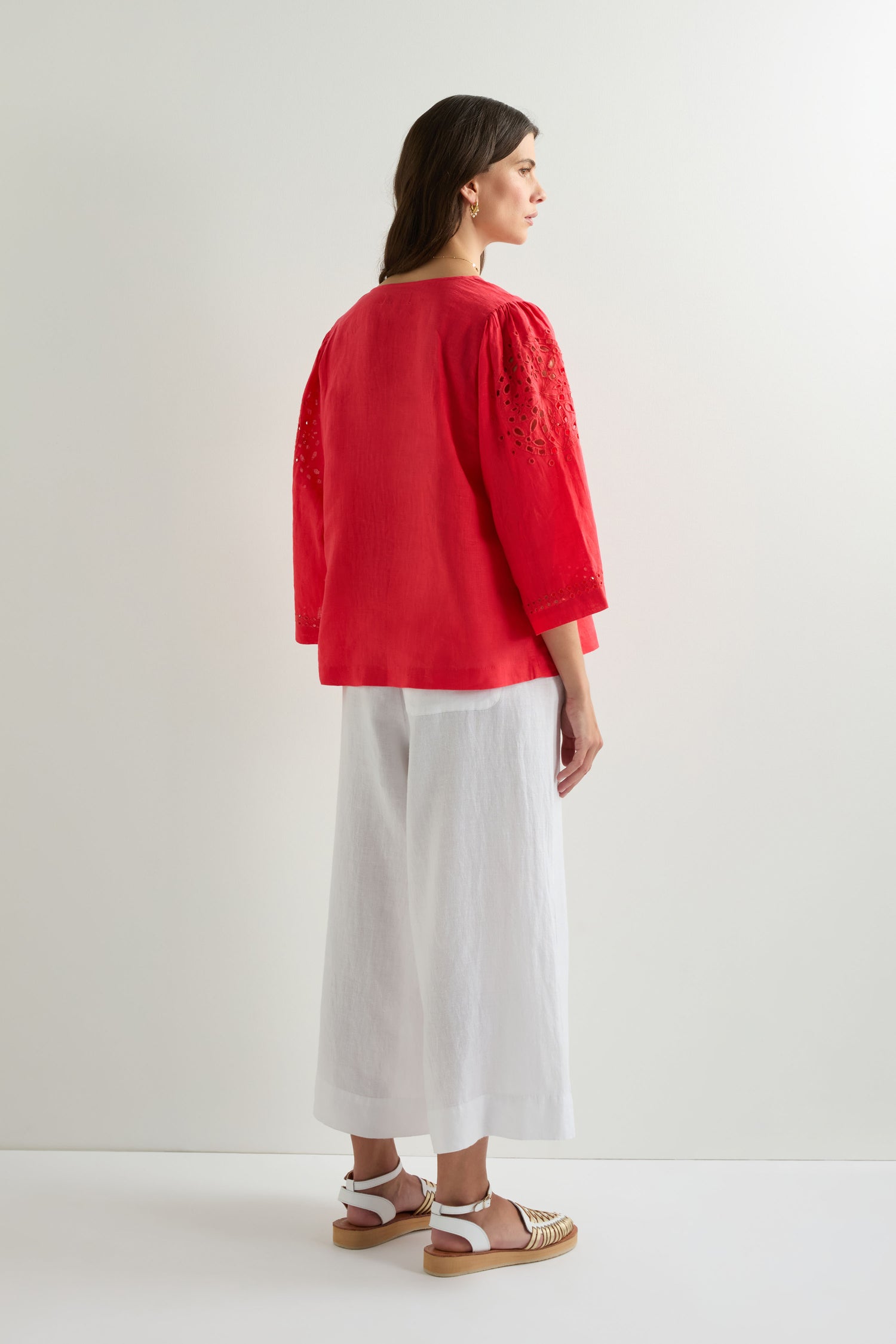 A woman faces away from the camera, wearing the Embroidered Schiffli Linen Top in red with 3/4 sleeves, paired with white wide-leg pants and sandals against a plain white background.