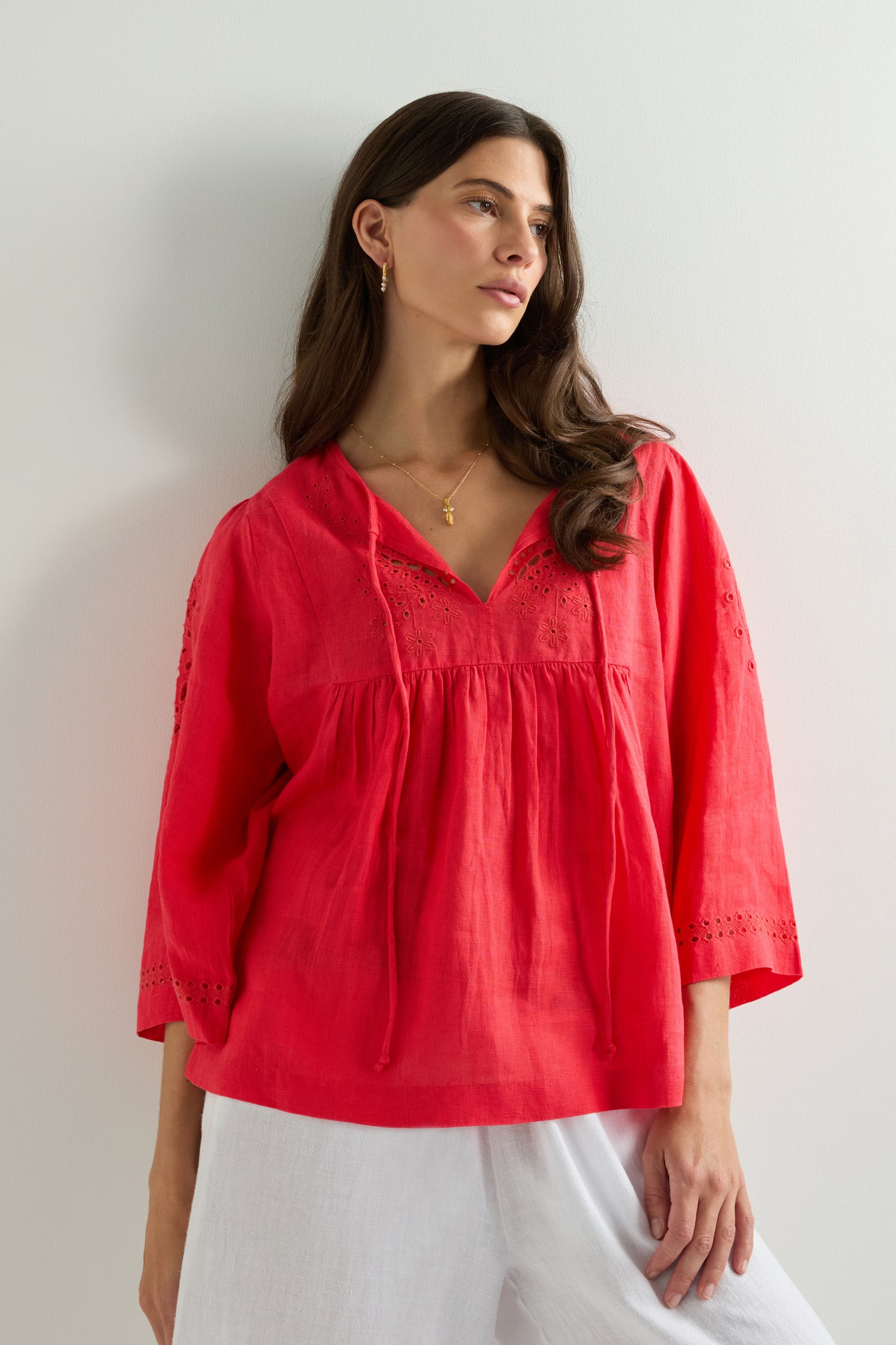 A woman wears the Embroidered Schiffli Linen Top, featuring lace-detailed sleeves, paired with white pants while standing against a plain light-colored wall.