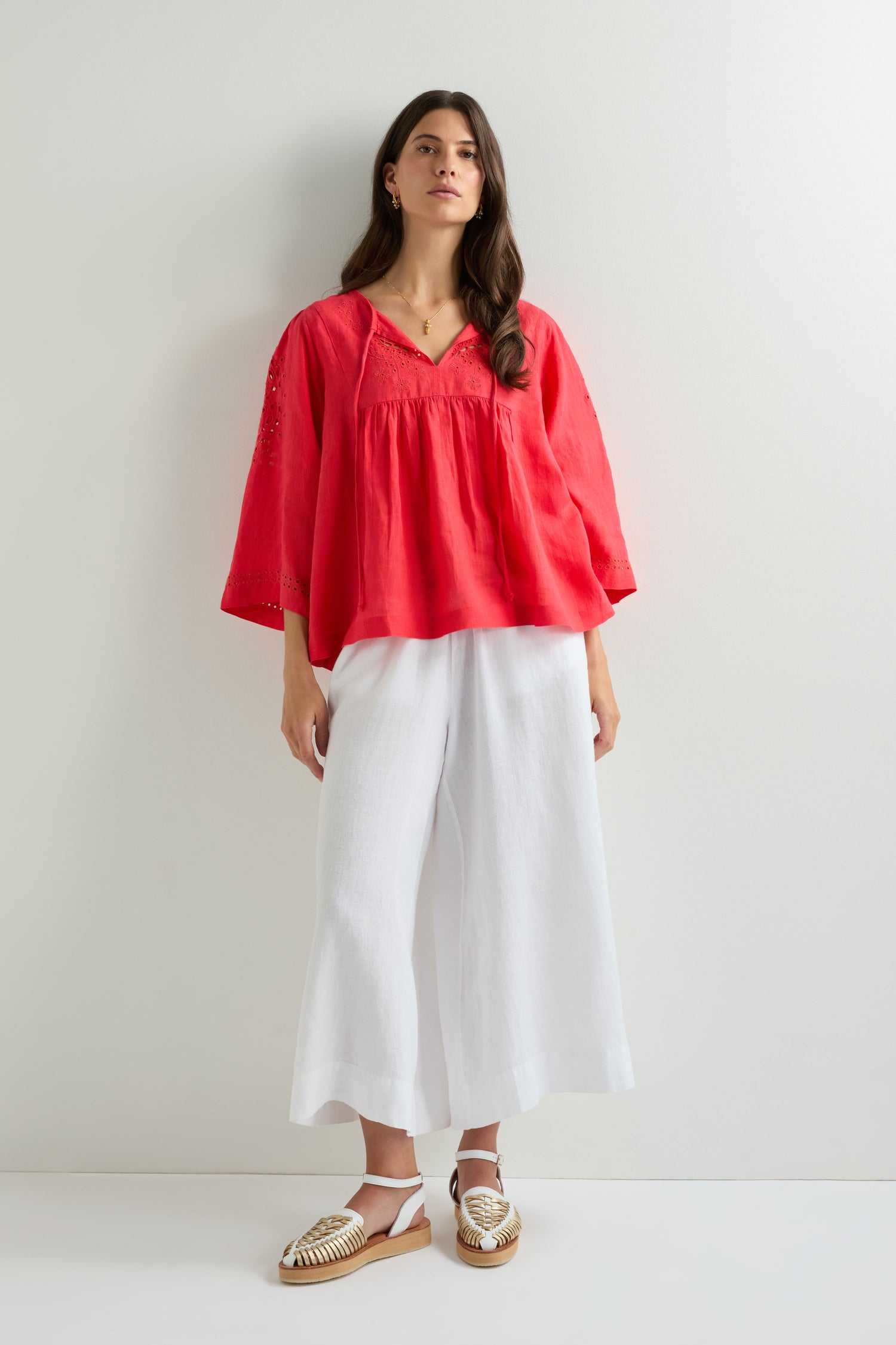 A woman stands against a white wall wearing an Embroidered Schiffli Linen Top in red, paired with wide white pants and white woven sandals.