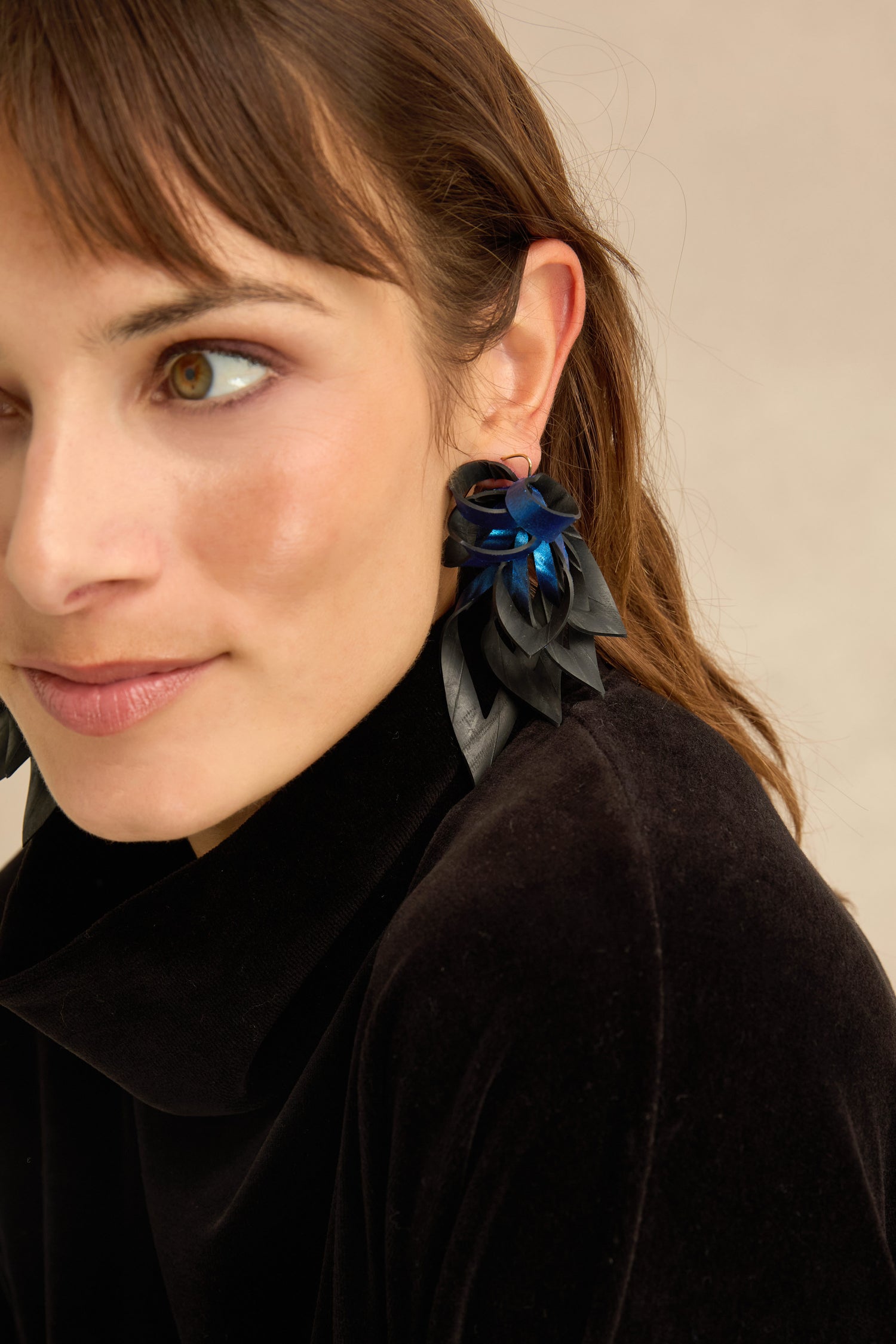 A woman wearing a black top and the Metallic Blue Flipped Leaves Earrings—large, flower-shaped accessories thoughtfully crafted from reclaimed bicycle inner tube in striking black and blue—looks slightly to the side.