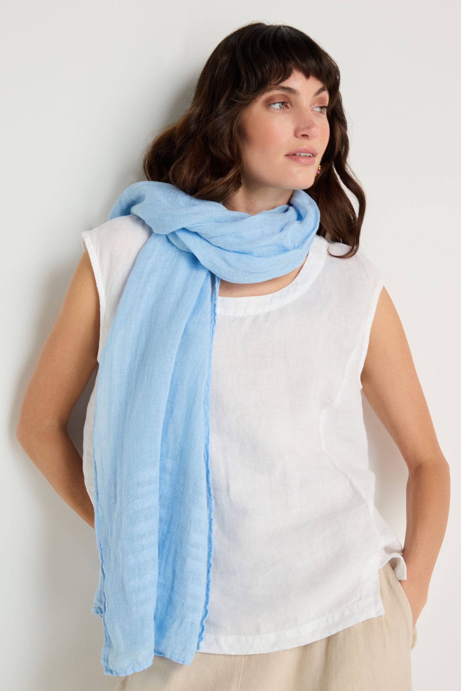 A woman in a sleeveless white top and beige pants stands against a white background, with a light blue Crinkle Linen Scarf draped around her neck and shoulder.