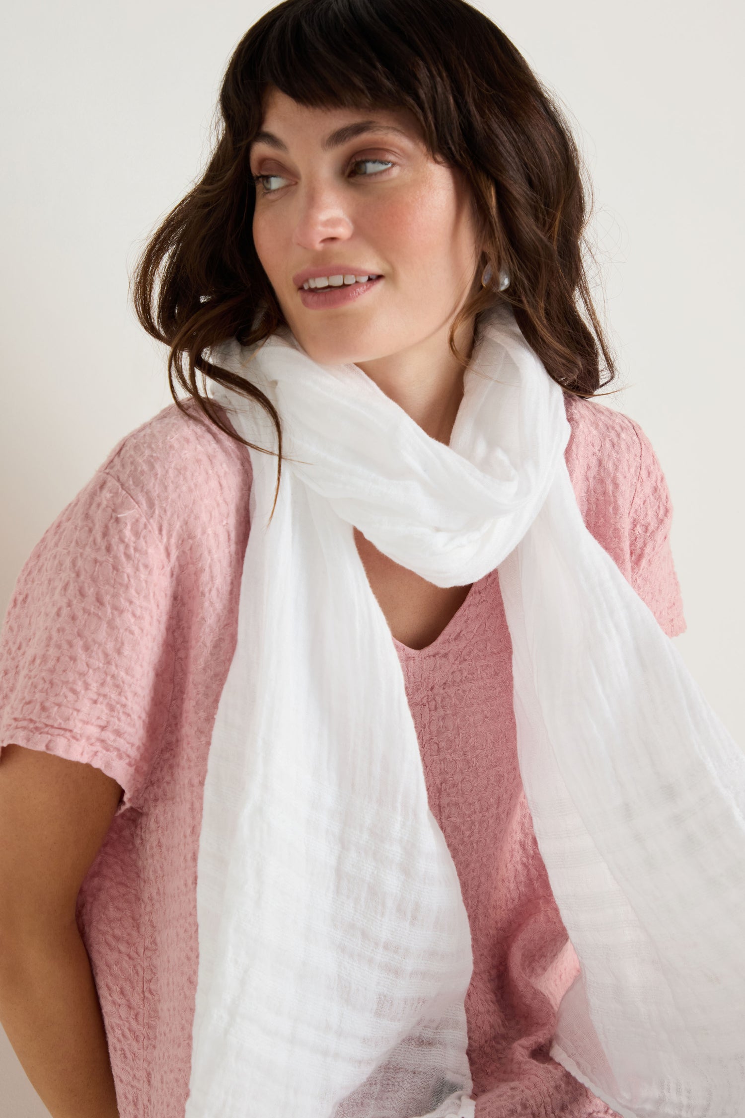 Woman with medium-length brown hair wears a textured pink top and the Crinkle Linen Scarf, looking slightly to the side with a neutral expression.