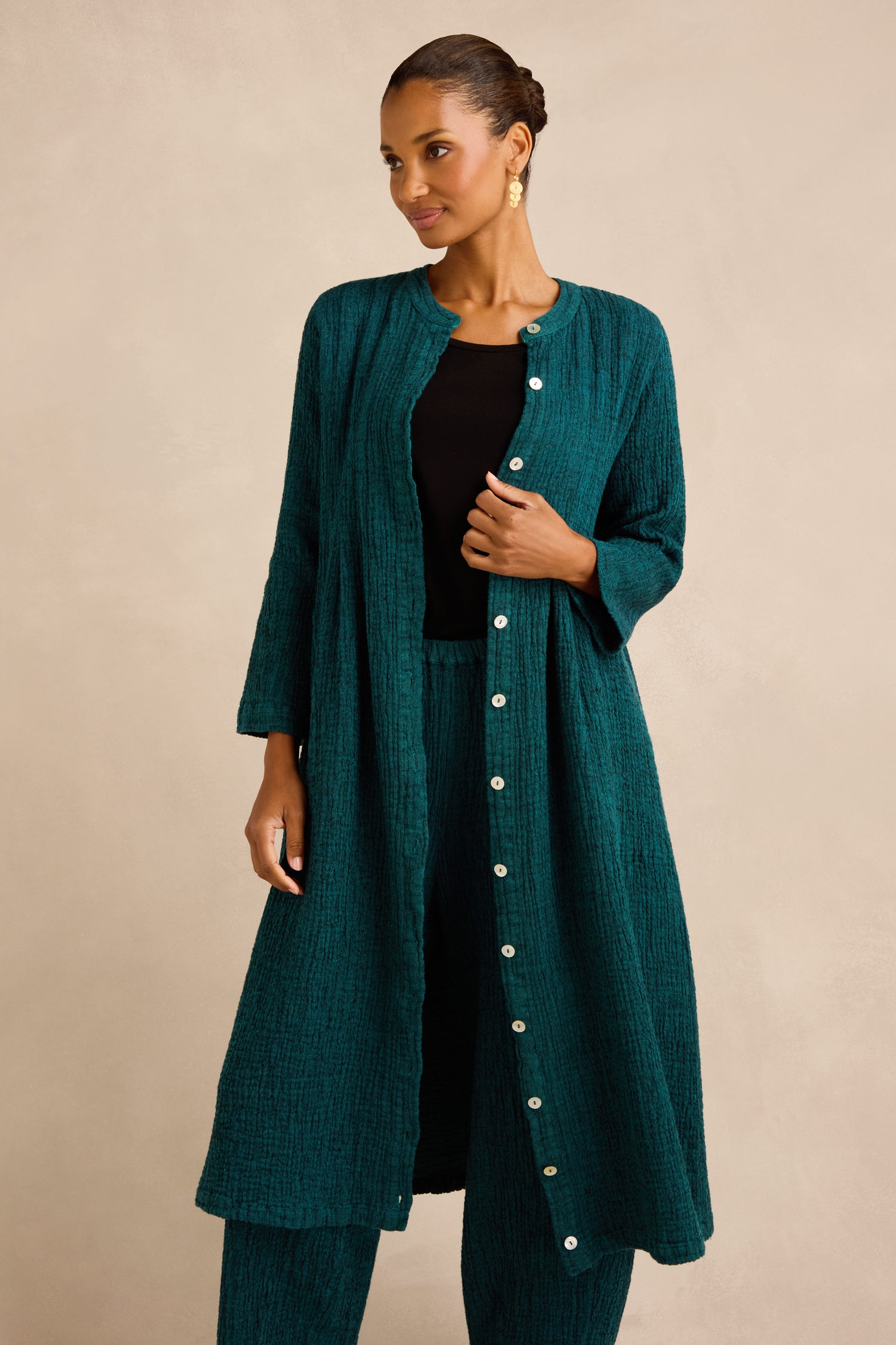 Woman wearing a teal Crinkle Double Face Linen Dress styled as a button-down long coat over matching teal pants and a black top, standing against a beige background. This dress adds effortless elegance with its unique crinkled texture and relaxed silhouette.