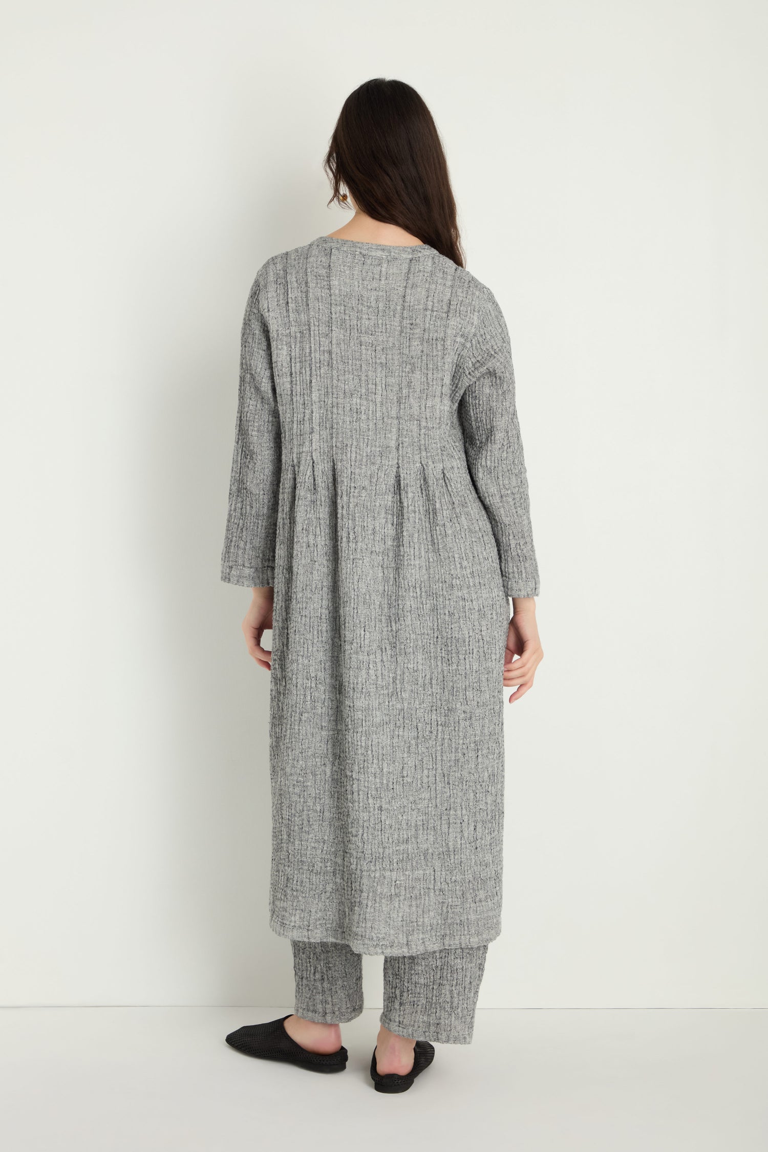 A person stands facing away, wearing the Crinkle Double Face Linen Shirt Dress in textured gray with matching pants and black slippers, set against a plain white background.