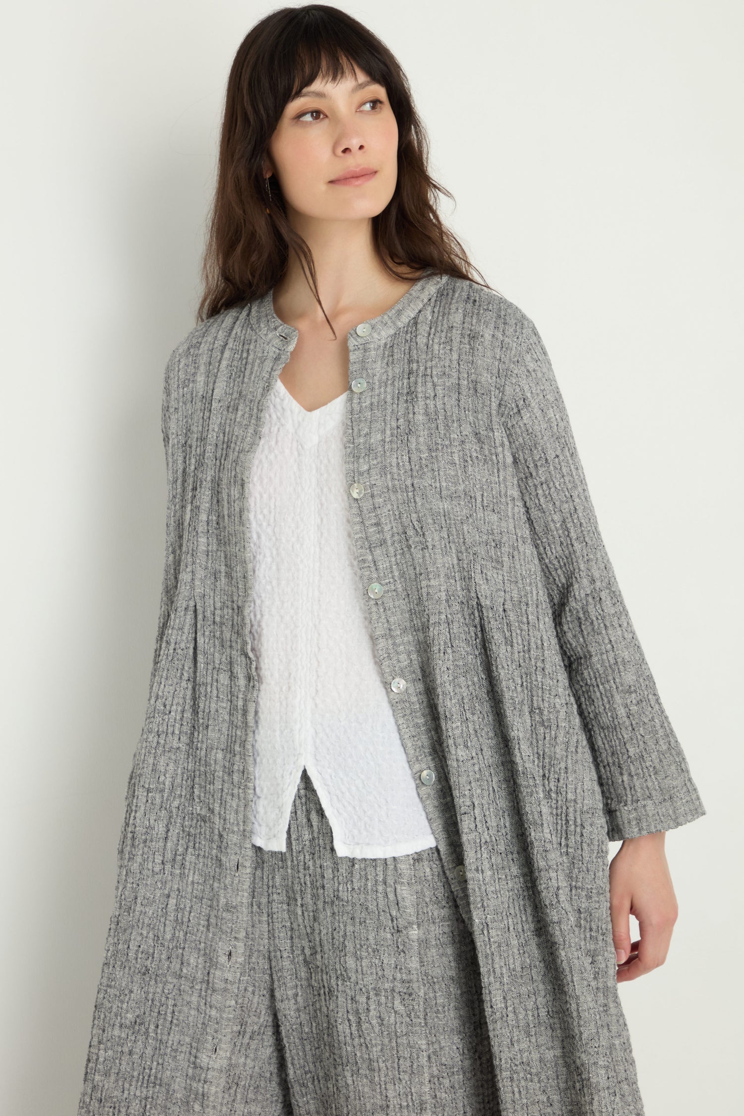 A woman stands against a plain light background wearing the Crinkle Double Face Linen Shirt Dress in textured grey over a white top and matching grey trousers.