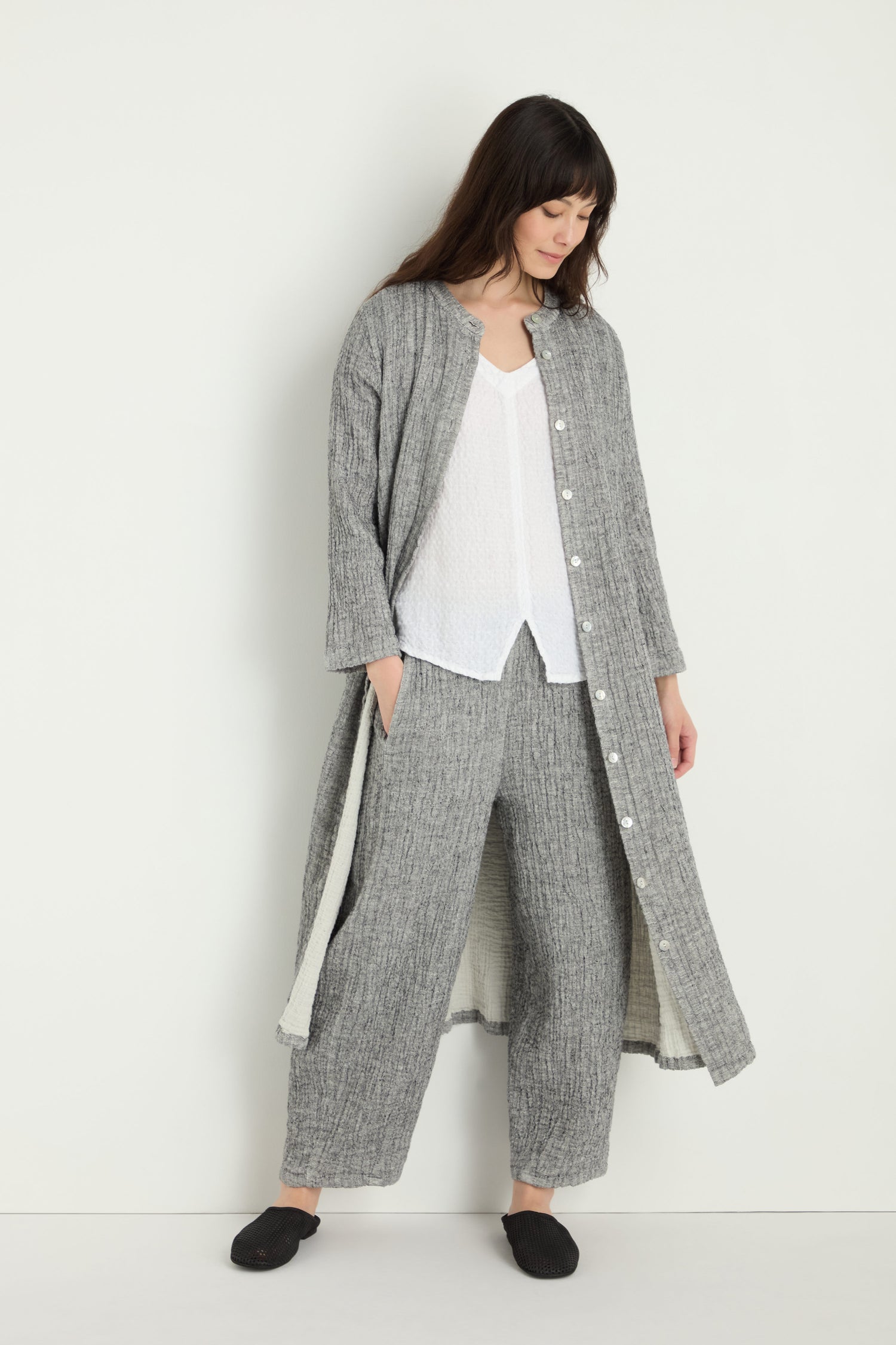 A woman stands against a white wall wearing the Crinkle Double Face Linen Shirt Dress in gray, styled as a long button-up coat, with matching wide-leg pants, a white top, and black slippers.