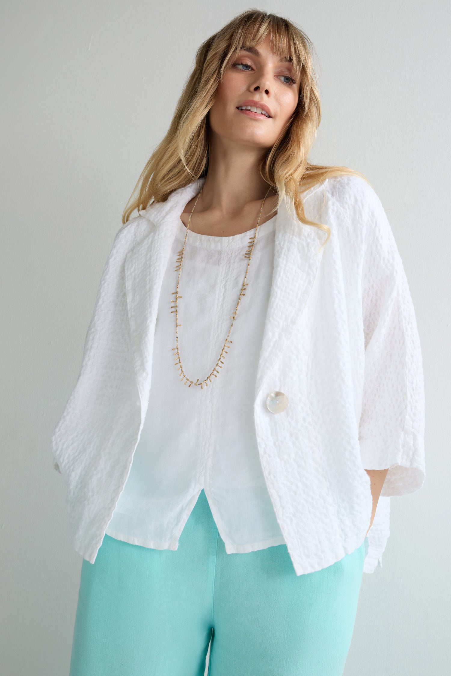 Woman wearing the Garment Dyed Waffle Linen Boxy Jacket over a white blouse with a long necklace and light blue pants, standing against a plain light-colored background.