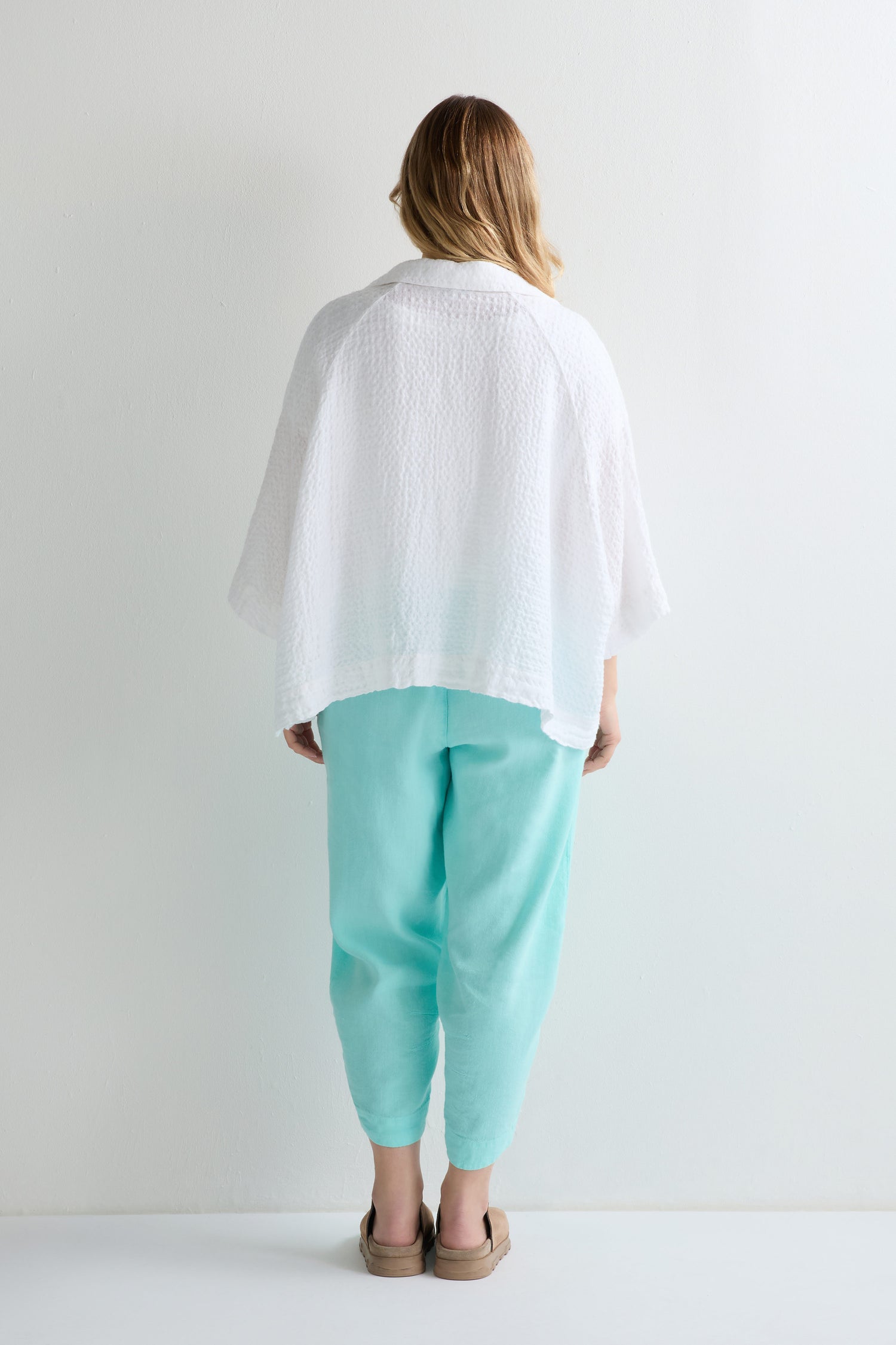 A person with shoulder-length hair, seen from behind and facing a white wall, wears the Garment Dyed Waffle Linen Boxy Jacket with an oversized fit, light blue pants, and beige sandals.