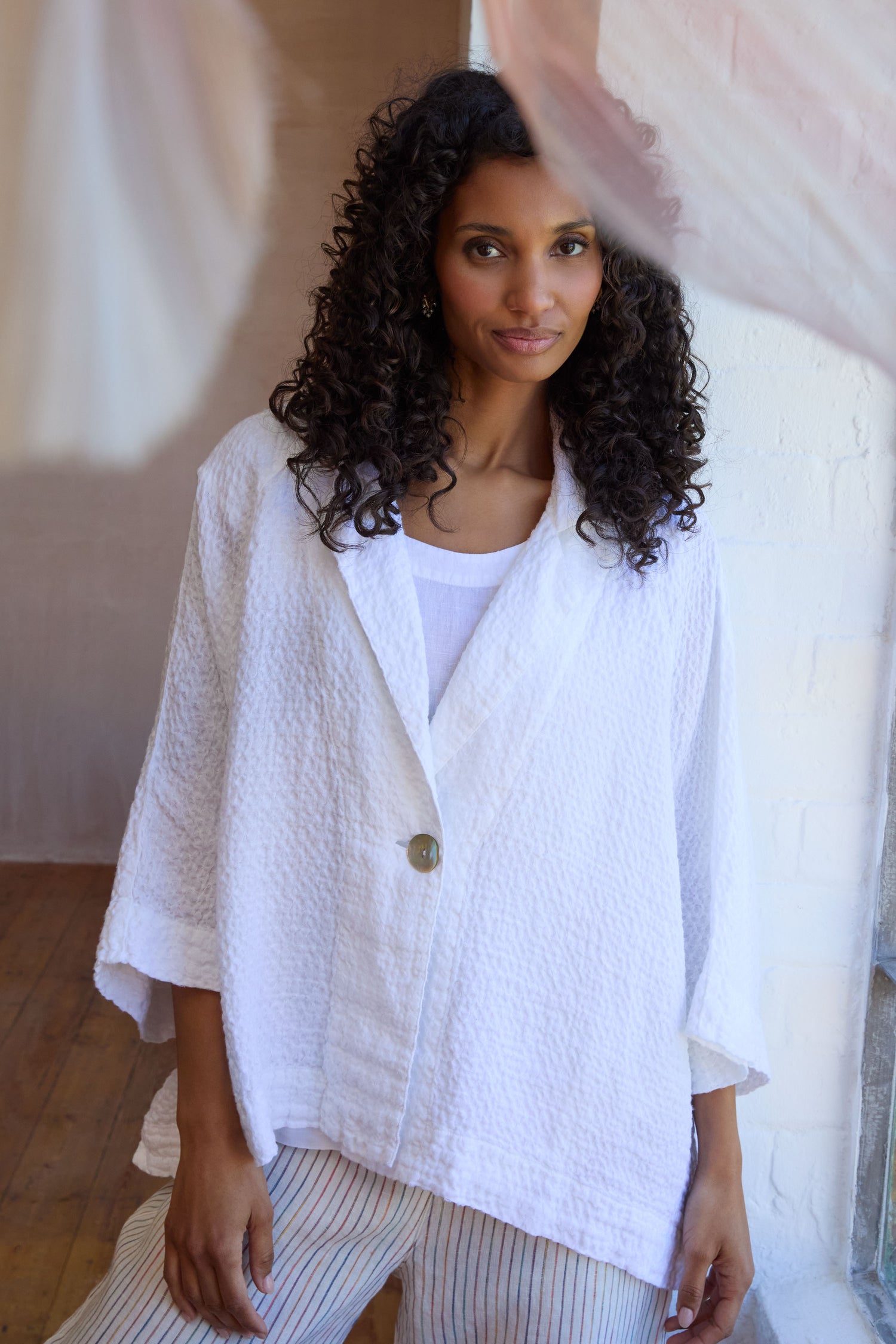 A woman with curly hair wears the Garment Dyed Waffle Linen Boxy Jacket over a white top and striped pants, standing indoors near a white brick wall.