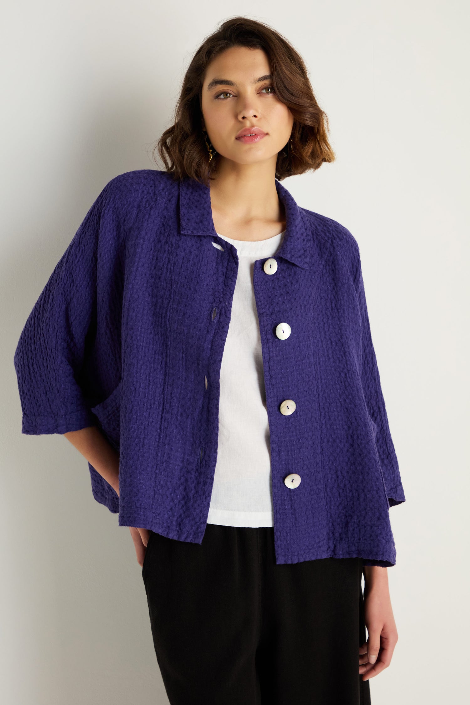 A woman stands against a light wall, wearing the Waffle Linen Boxy Jacket in purple over a white top and black pants.