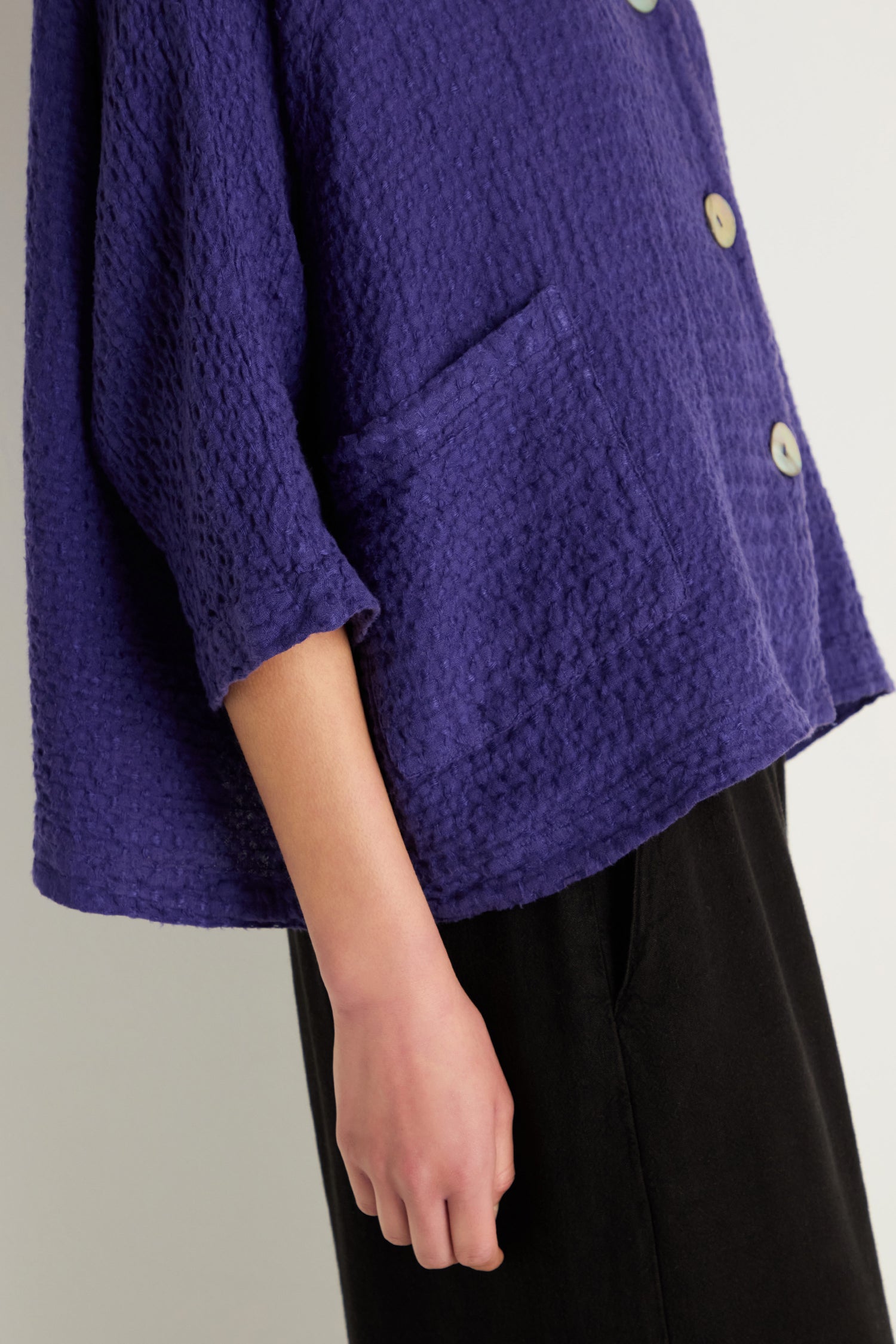 A person wearing the Waffle Linen Boxy Jacket in textured purple with a front pocket, paired with black pants, stands against a neutral background.