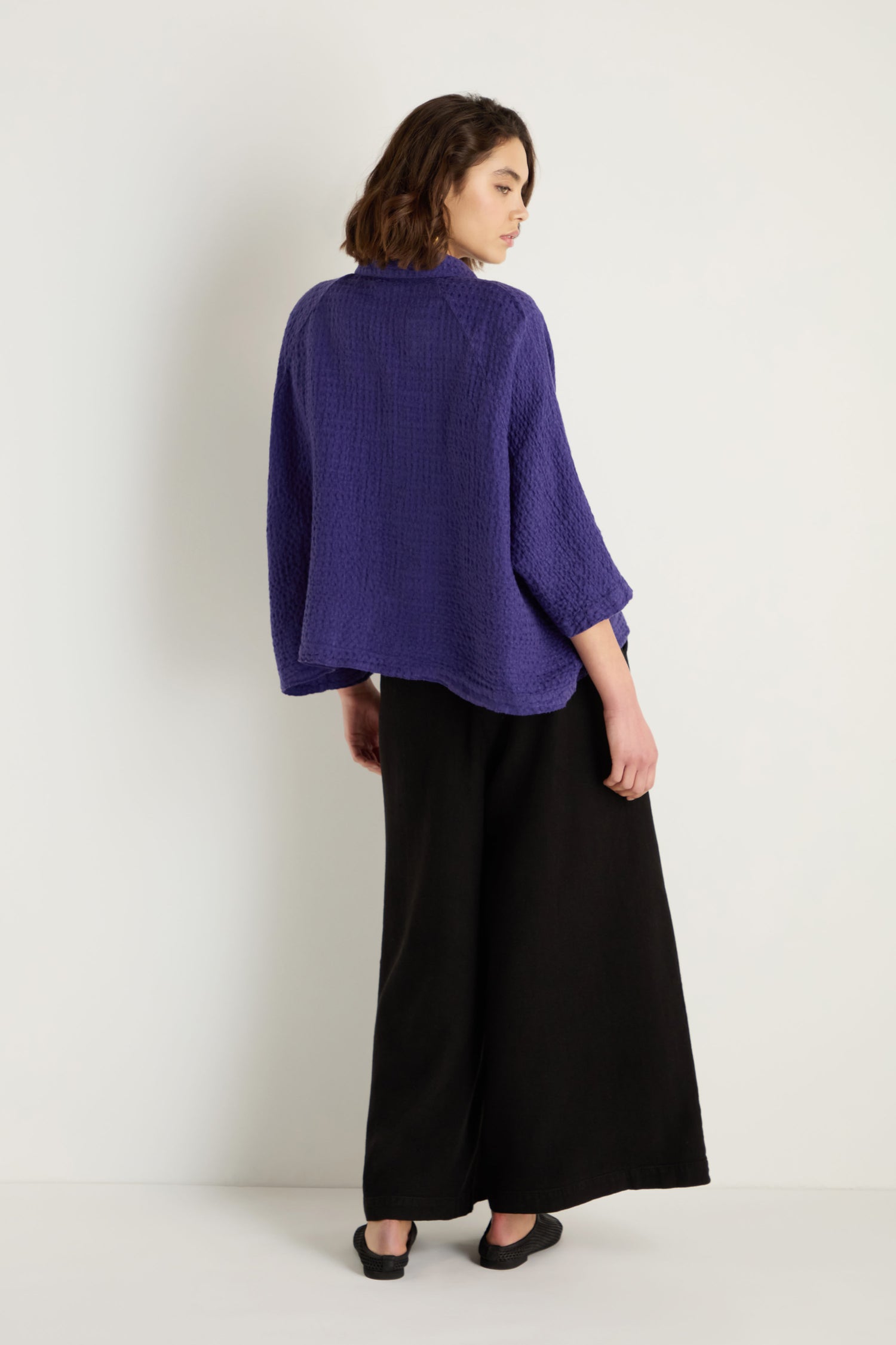 A woman stands slightly turned, wearing the Waffle Linen Boxy Jacket over a textured purple top, paired with wide-leg black pants and black shoes against a plain white background.