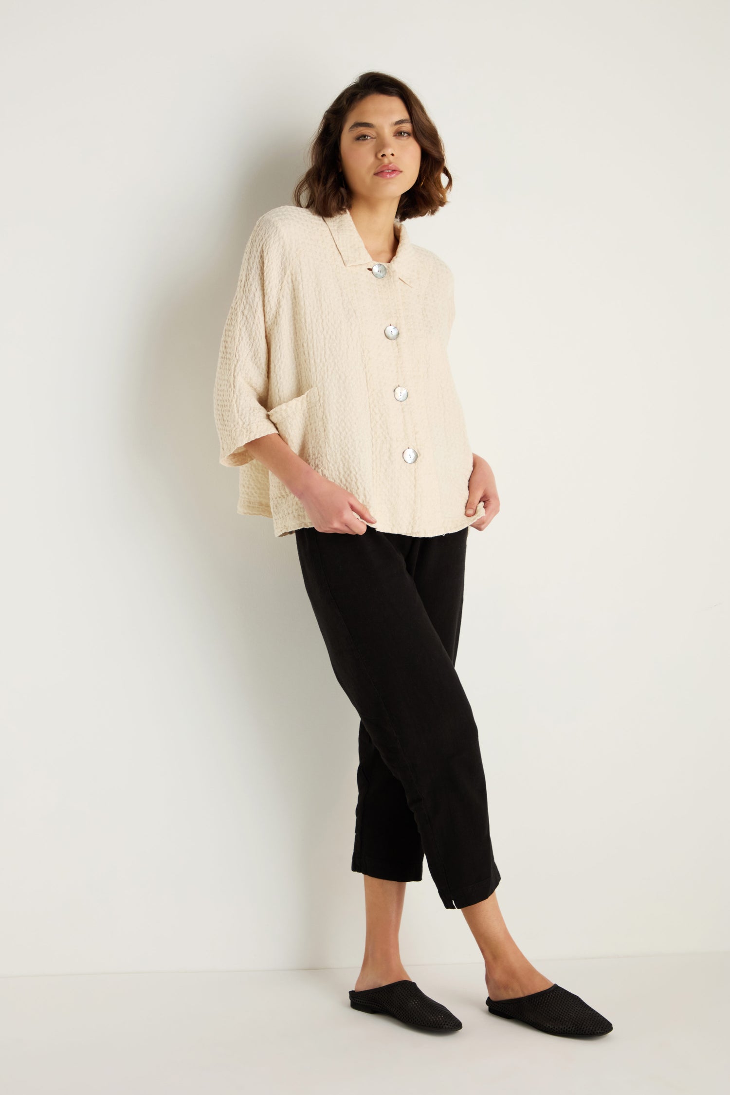 A person stands against a white wall wearing a Waffle Linen Boxy Jacket in textured cream with large buttons, paired with black cropped pants and black slip-on shoes.