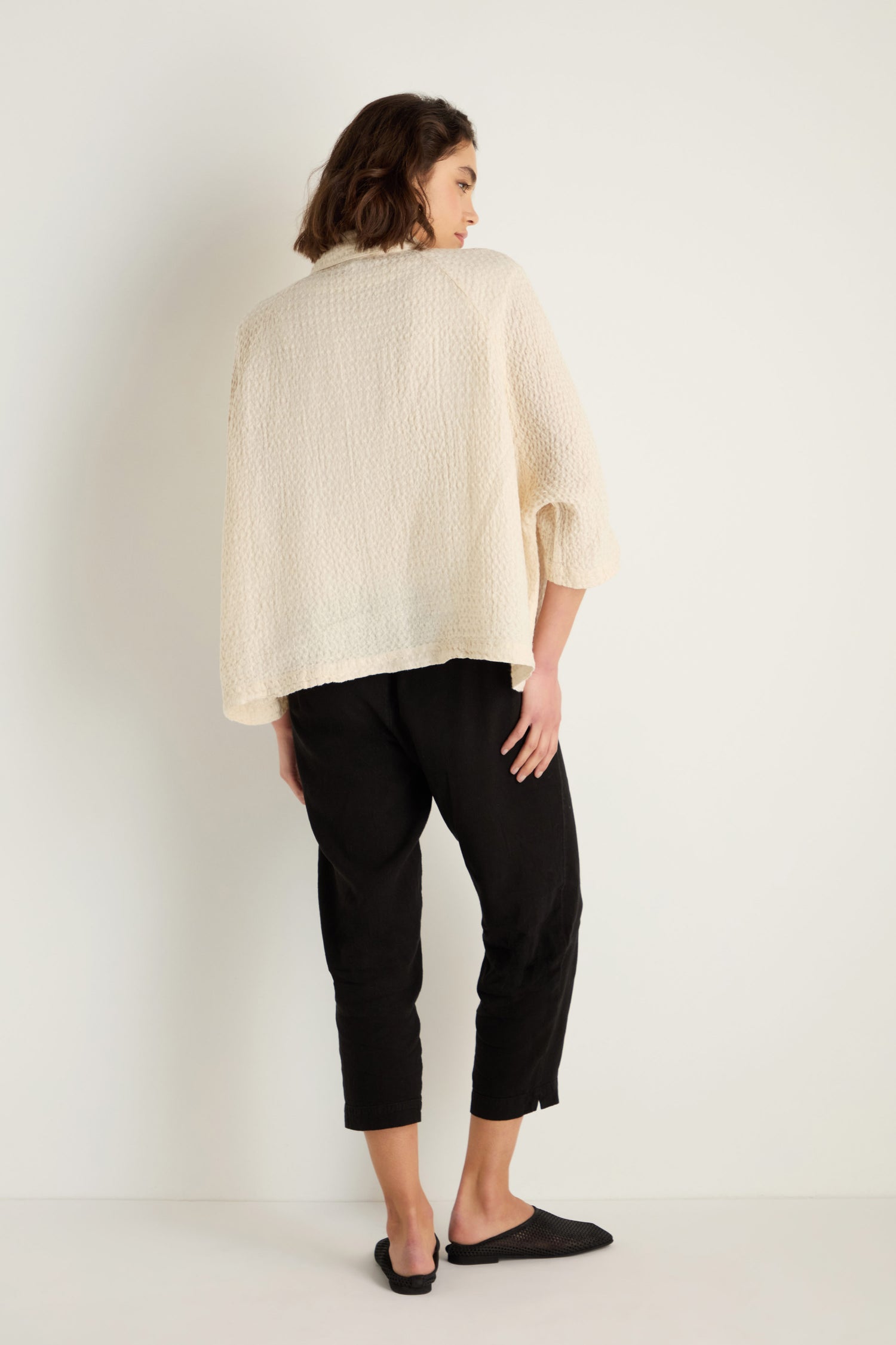 A person stands facing away from the camera, wearing a textured cream Waffle Linen Boxy Jacket, black cropped pants, and black flats against a plain white background.