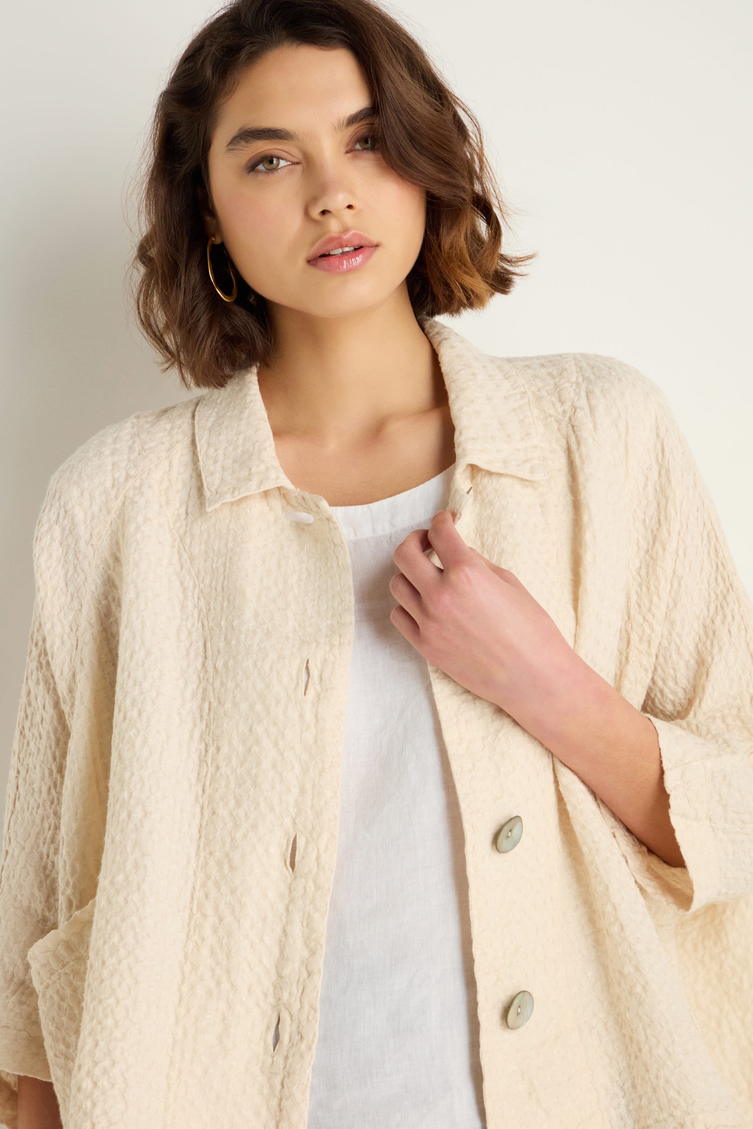 A woman with wavy brown hair wears the Waffle Linen Boxy Jacket in cream over a white top, standing against a plain light background.