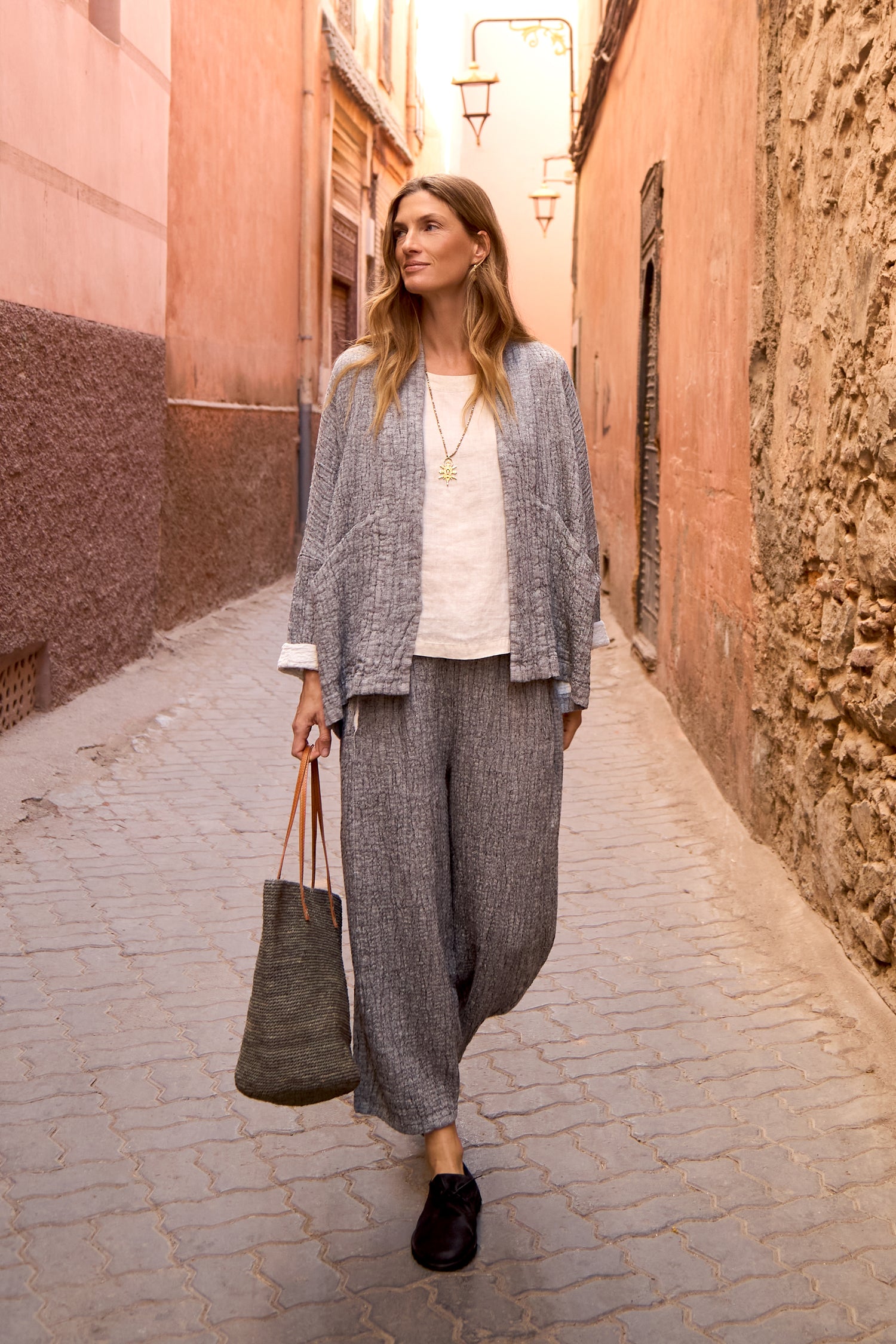 A woman strolls through a narrow, sunlit alley in loose grey clothes and a white top, carrying a large tote. She completes her look with the Crinkle Double Face Linen Jacket, effortlessly blending style and function.
