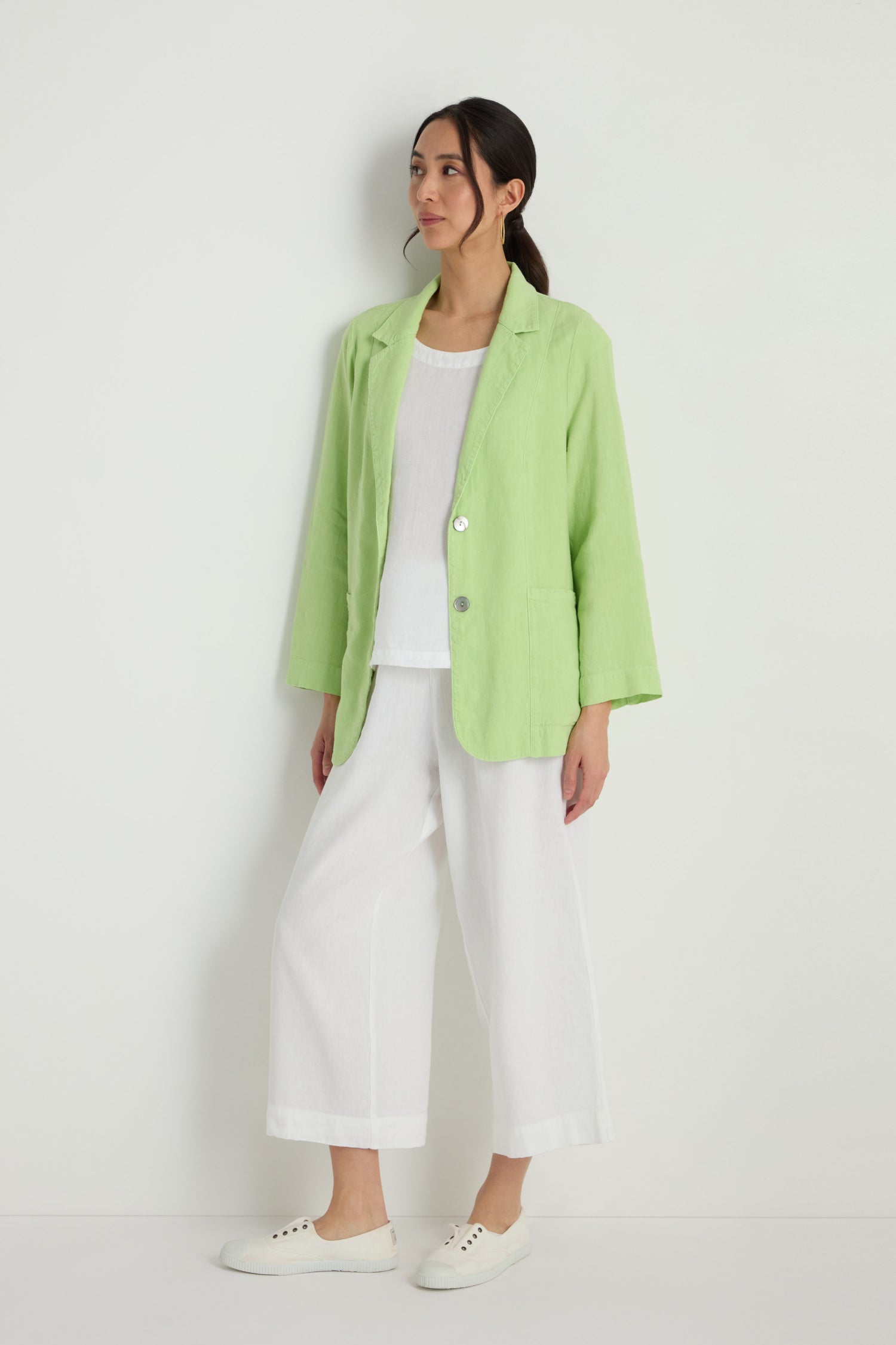 Person standing against a white wall wearing a Twisted Linen Relaxed Blazer in light green, paired with a white top, white wide-leg pants, and white sneakers.