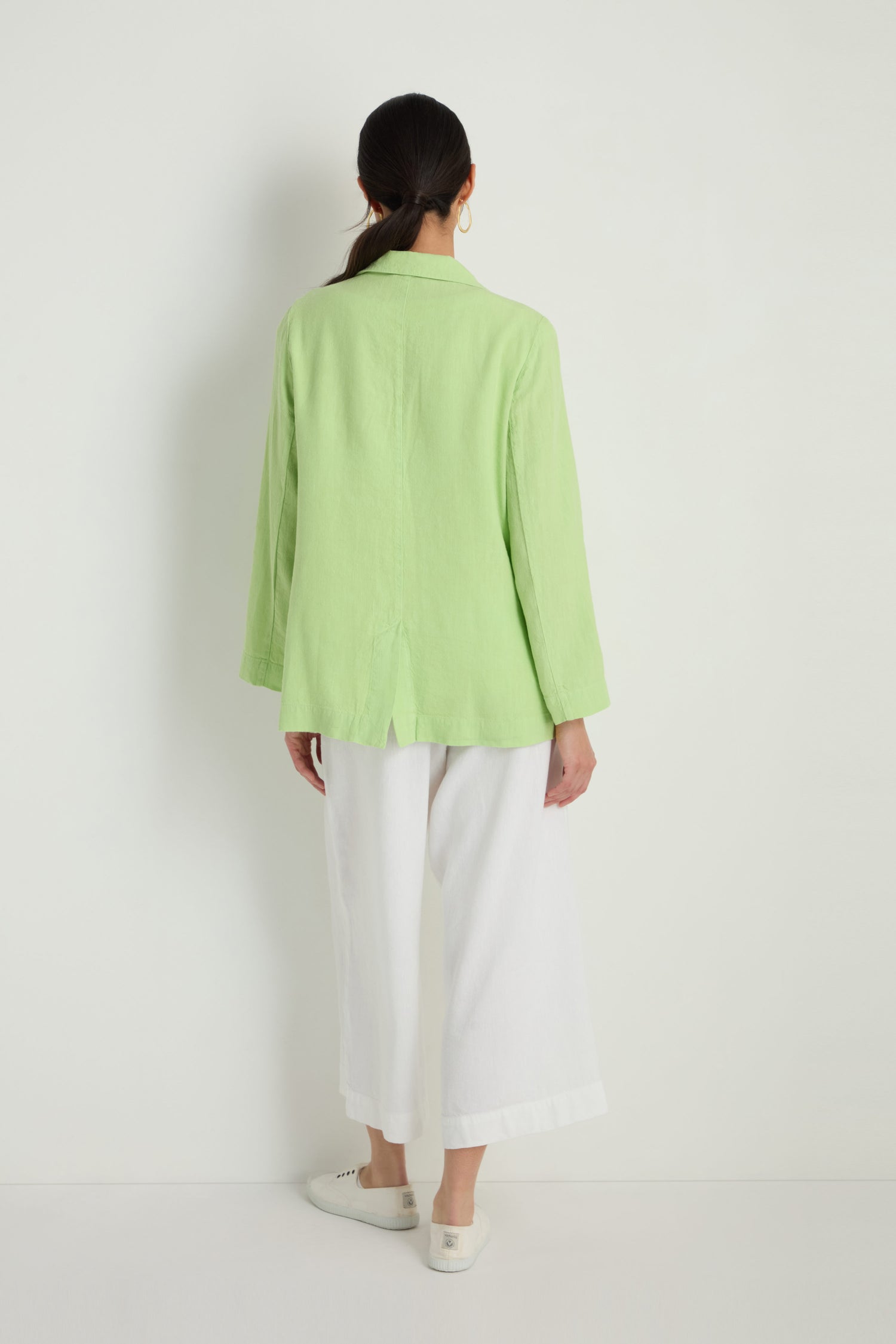 A person faces away, wearing the Twisted Linen Relaxed Blazer in light green, paired with white wide-leg pants and white shoes against a plain white background.