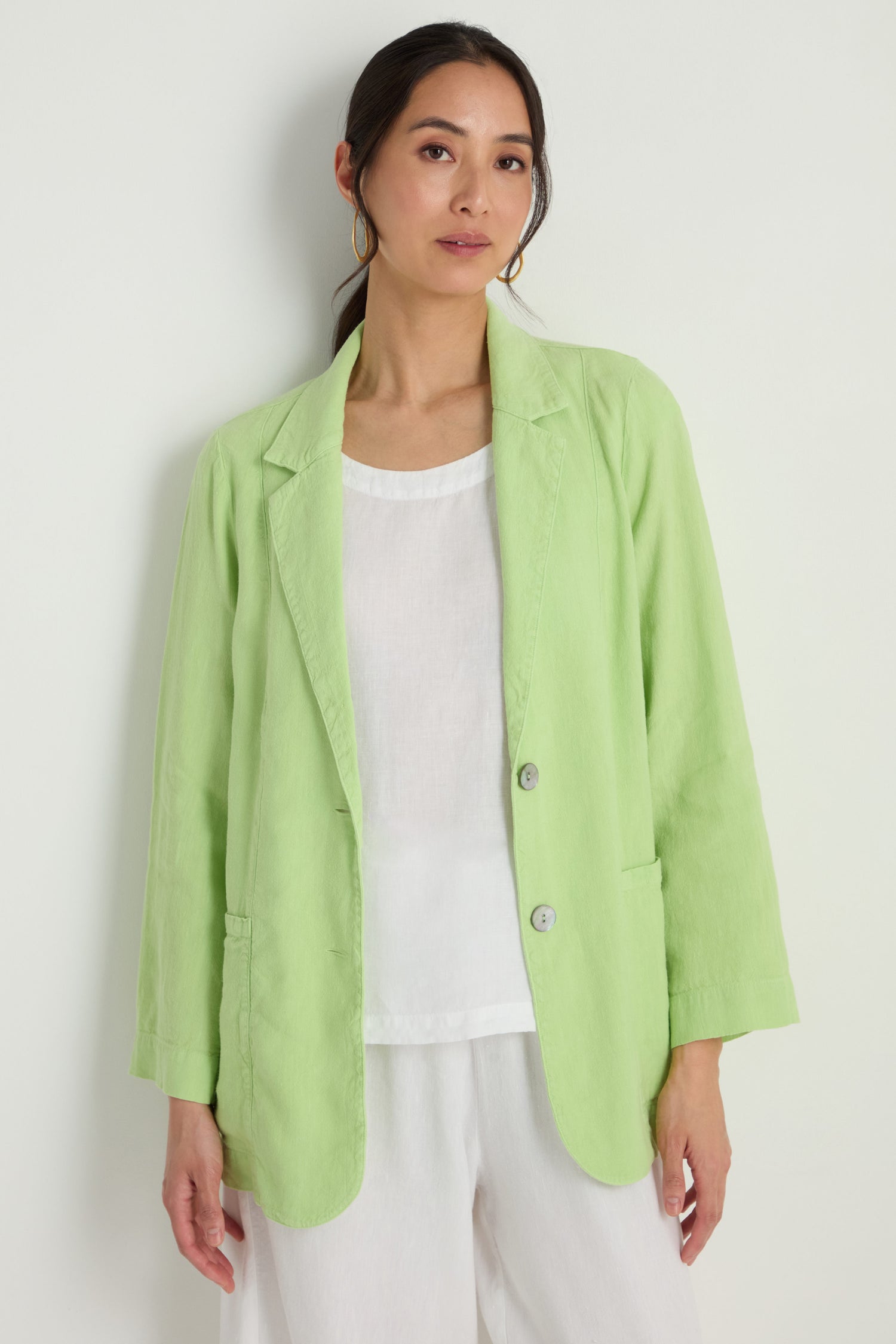 Model wears the Twisted Linen Relaxed Blazer in light green over a white top and white pants, standing against a plain light-colored background.