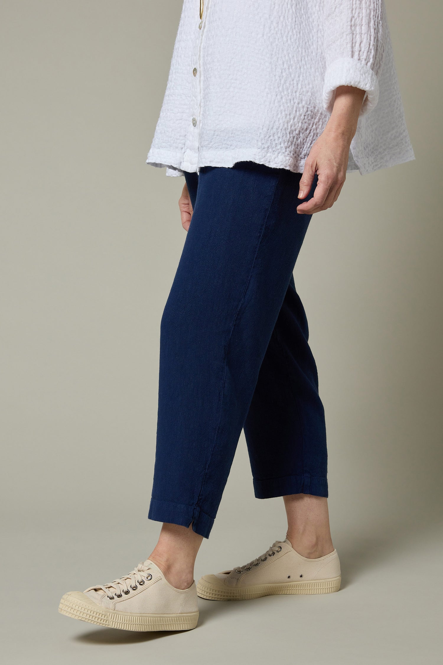 A person stands against a neutral background wearing a white textured long-sleeve shirt, Twisted Linen Navy Slim Trousers with an elasticated waistband, and beige lace-up sneakers.
