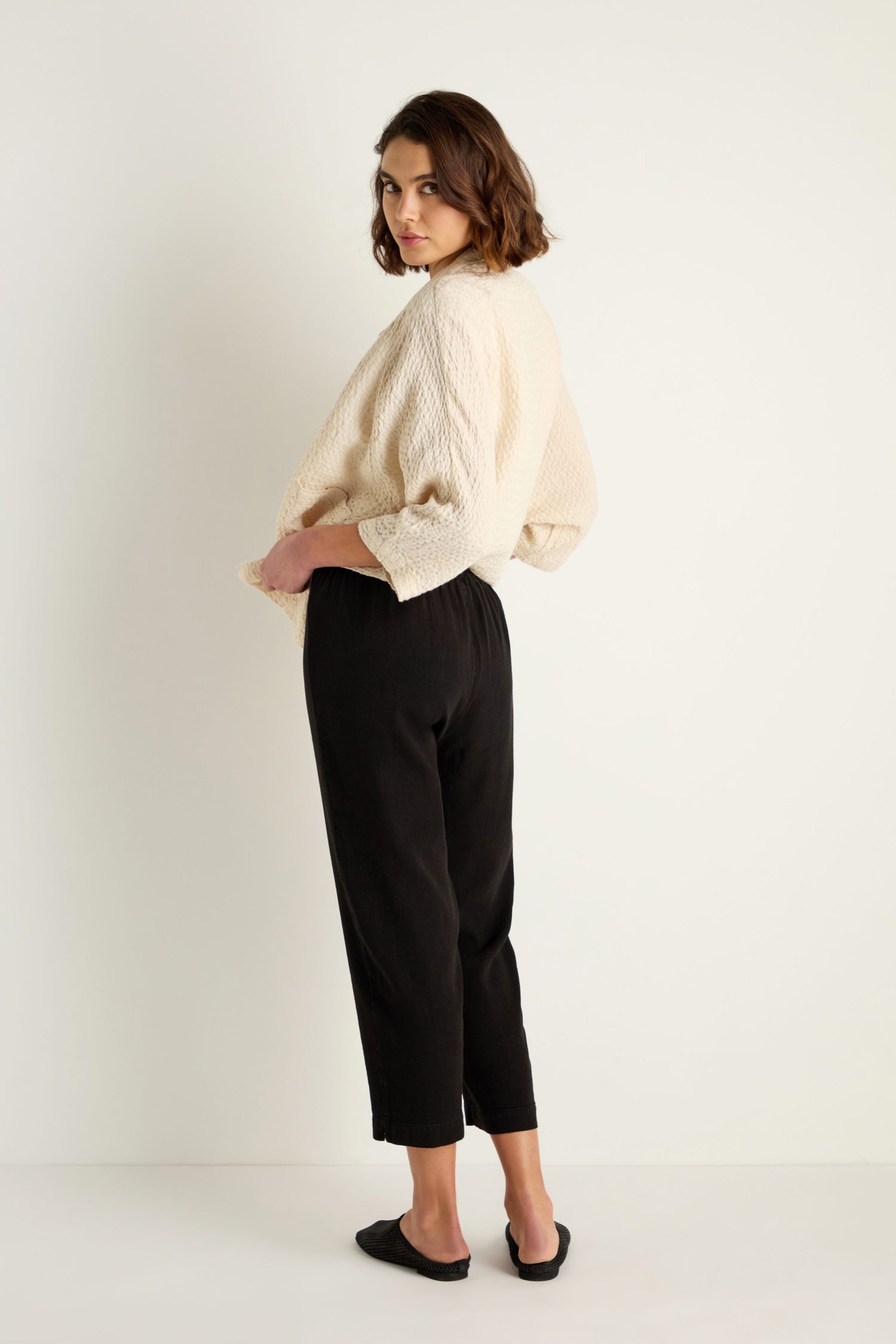A woman stands in a neutral setting, wearing a textured beige top, the Twisted Linen Slim Trouser in black, and black flats, looking over her shoulder at the camera.